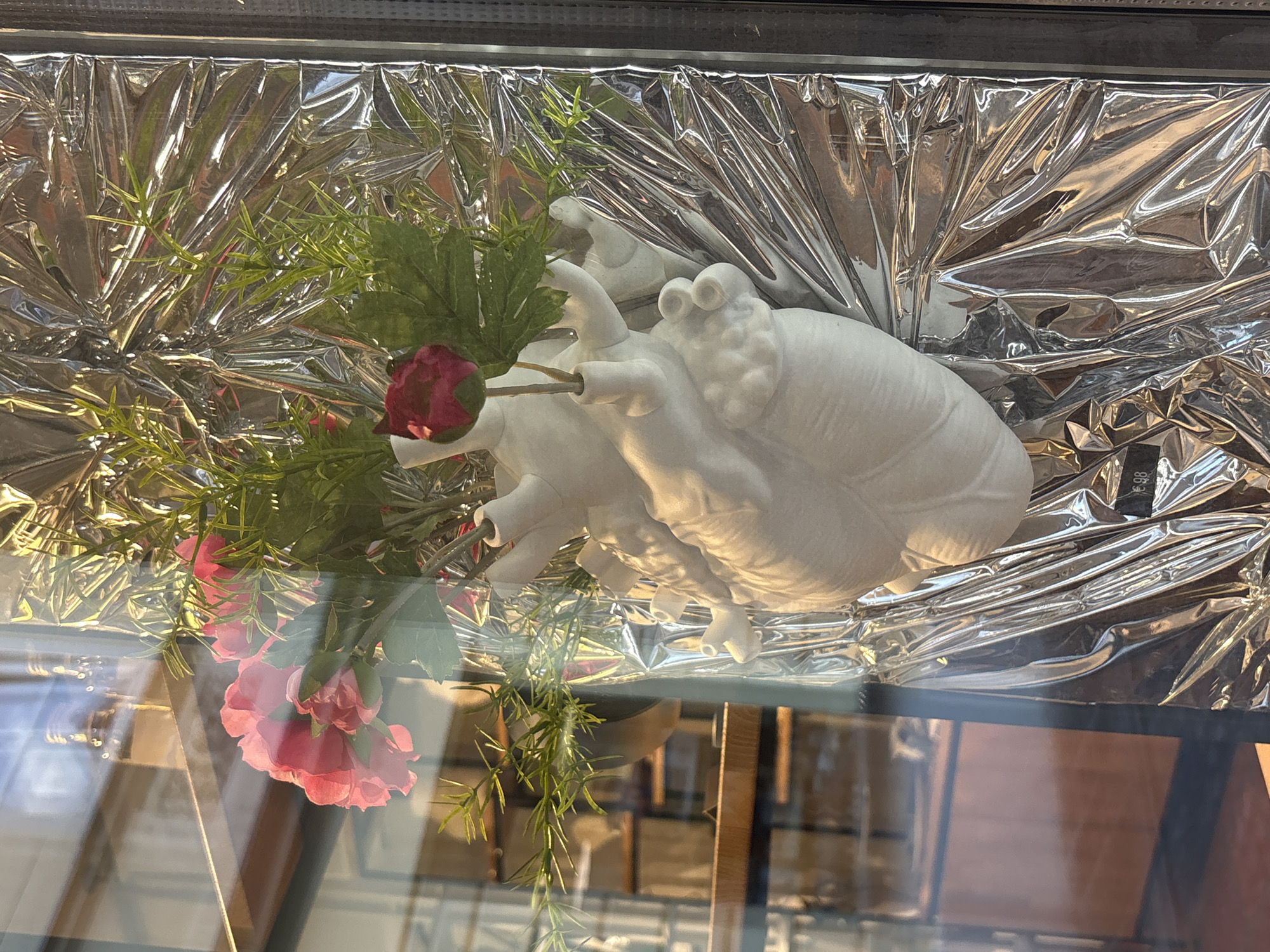 A white, anatomical heart-shaped vase holds a small bouquet of pink and red flowers and green sprigs. The vase is positioned in front of a wall covered with crinkled, reflective silver material. The scene is viewed through a glass pane, which shows reflections of shelves and possibly bottles in the background.