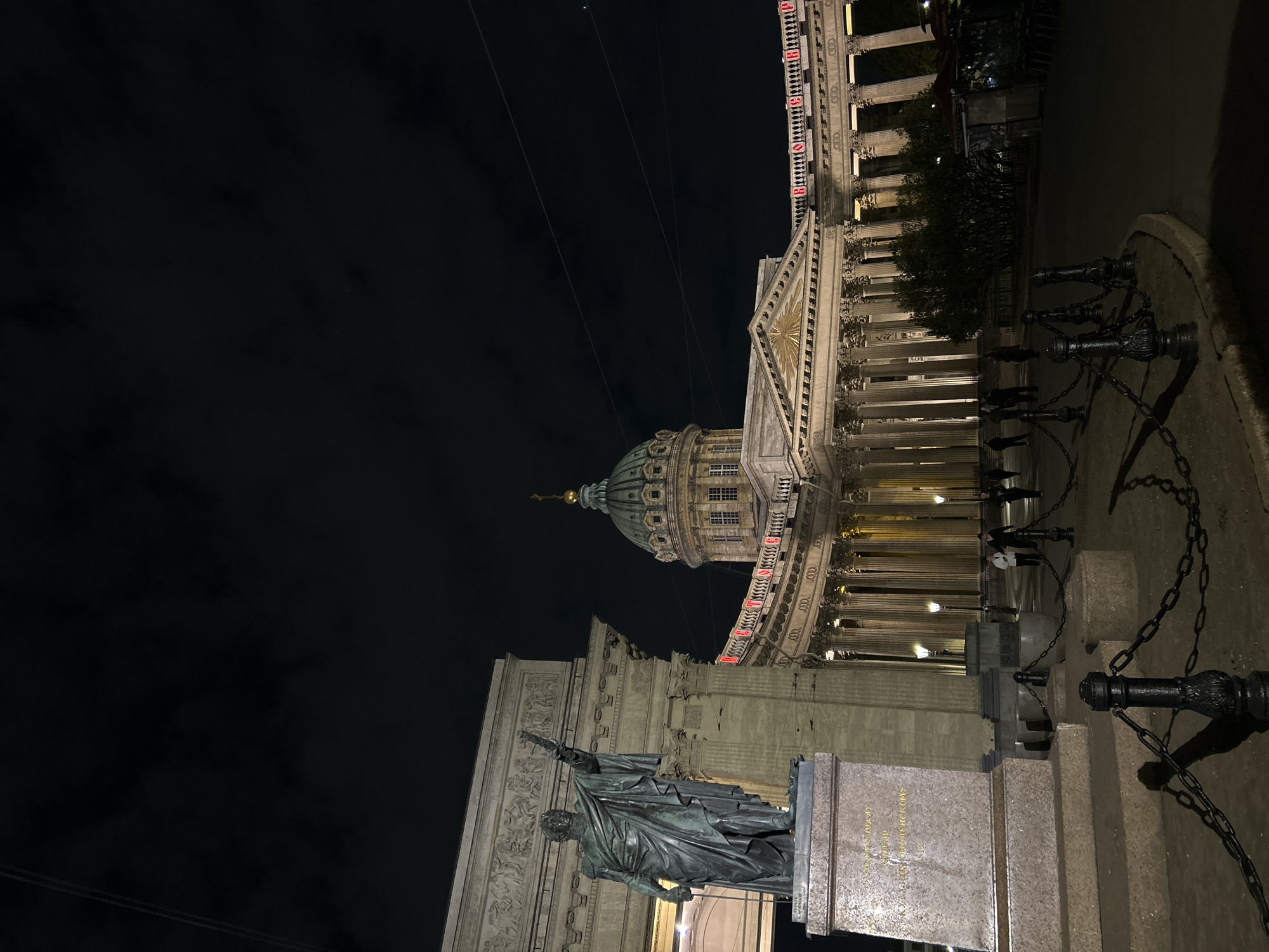 The image shows Kazan Cathedral and the monument to Michael Barclay de Tolly in St. Petersburg, Russia, at night. The cathedral's dome and colonnades are visible, illuminated by lights. A statue of Barclay de Tolly stands in the foreground. The sky is dark, and some people are walking in the background. A chain fence runs along the sidewalk in the foreground.