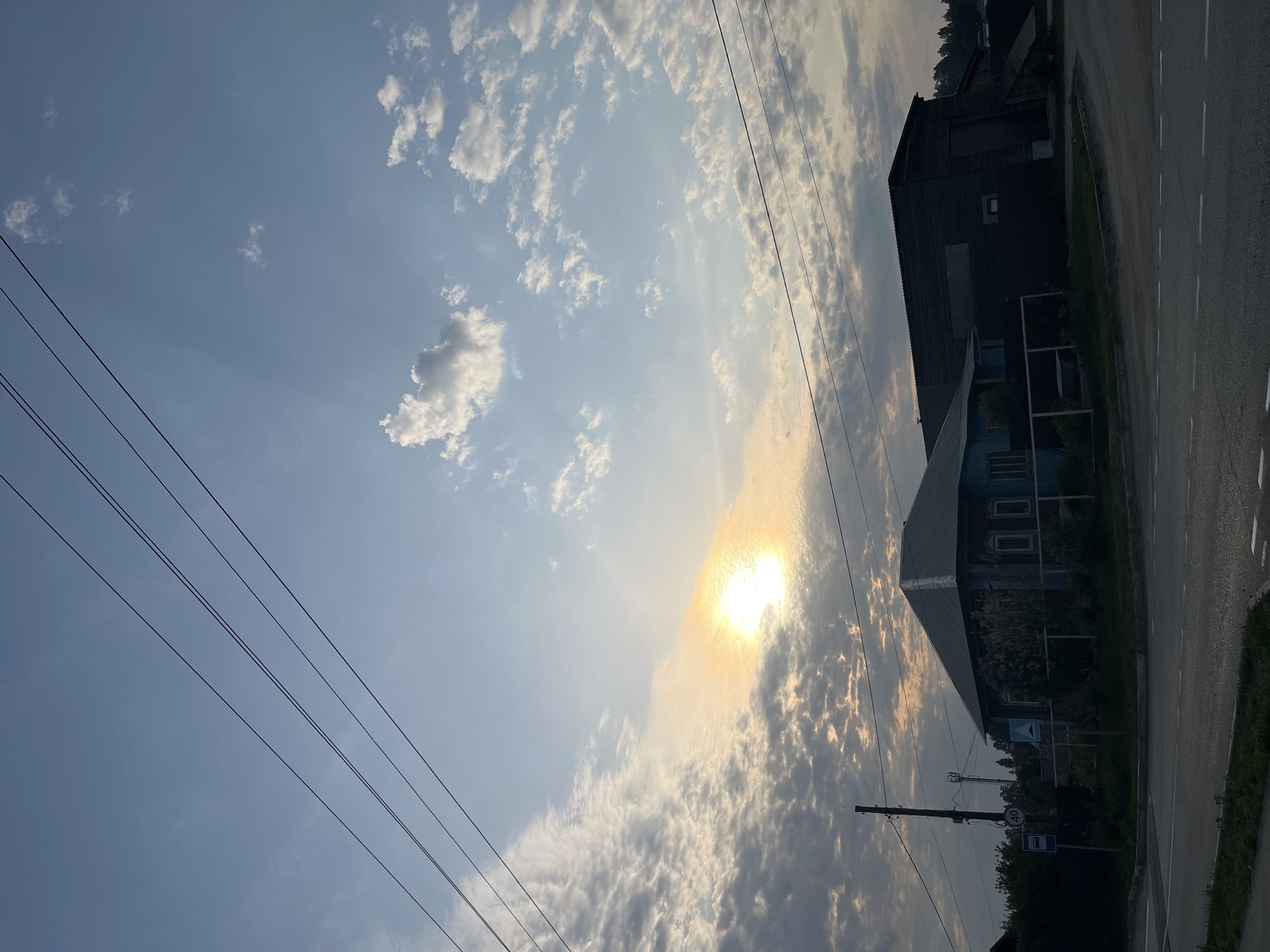 The image shows a daytime scene in Chaikovsky, Russia. Overhead, a bright sun is partially obscured by a thick layer of clouds. Power lines stretch across the left side of the frame. To the right, there is a road bordered by a building, foliage, and a bus stop sign. The image is rotated, and the elements appear to be tilted to the right.