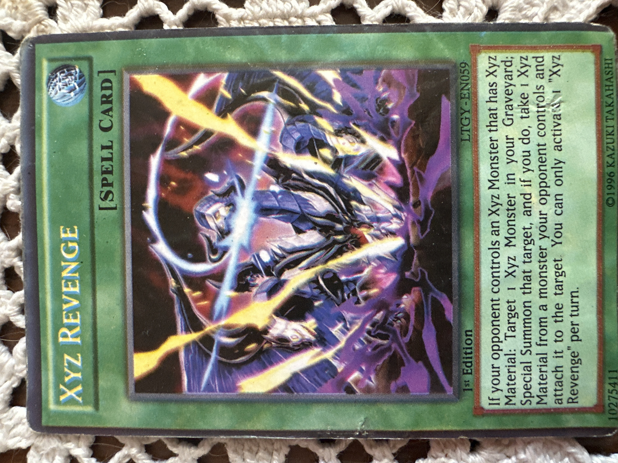 The image shows a Yu-Gi-Oh! trading card named "Xyz Revenge". It is a green "Spell Card". The card's text describes its effect: if the opponent controls an Xyz Monster with Xyz Material, the player can target an Xyz Monster in their Graveyard, Special Summon it, and attach an Xyz Material from the opponent's monster to the Special Summoned monster. The card's set code is LTGY-EN059, and it is a 1st Edition card. The bottom right corner displays the copyright information of ©1996 Kazuki Takahashi and the card ID 10275411. The card is lying on a crocheted surface.