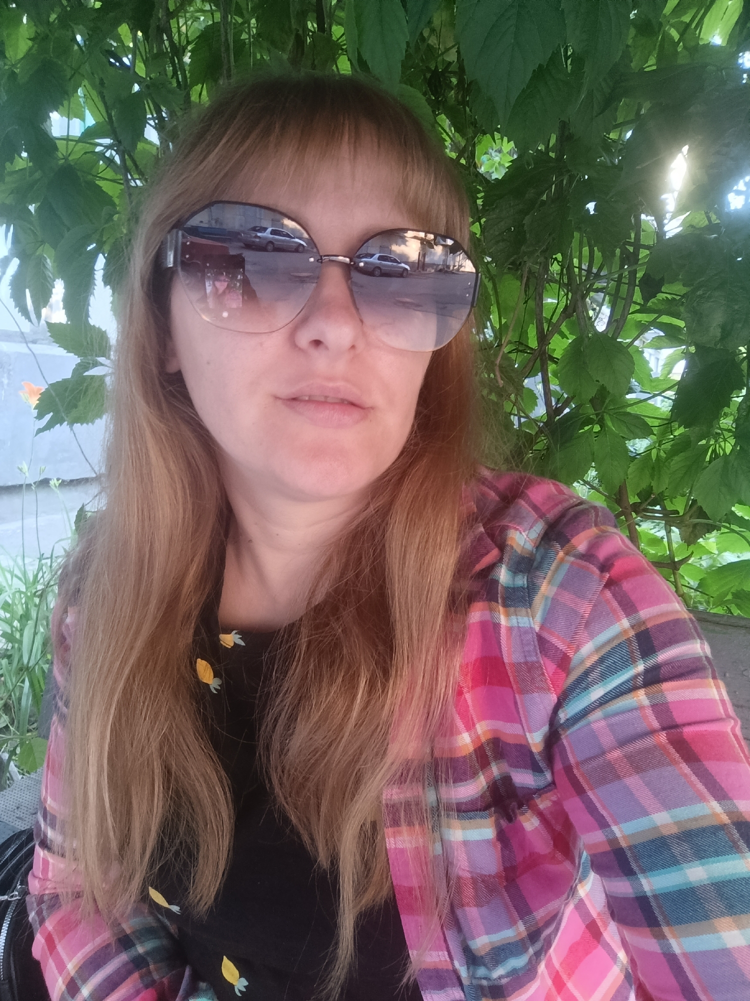 The image is a medium shot selfie of a woman with blonde hair and bangs, wearing large, round sunglasses and a plaid shirt. She is outdoors, with lush green foliage behind her, suggesting a daytime setting with sunlight filtering through the leaves. The woman's face is partially obscured by her sunglasses, but her mouth is slightly open, and her expression appears neutral or perhaps slightly amused.
The sunglasses reflect a street scene with cars and buildings, indicating an urban or suburban environment. Her shirt is a vibrant pink and purple plaid pattern. Underneath the unbuttoned shirt, she wears a black top adorned with small, yellow, carrot-like designs. Her long blonde hair falls over her shoulders.
The overall mood of the image is casual and relaxed, hinting at a warm and pleasant day. The background foliage provides a natural element, contrasting with the urban reflection in her sunglasses. There is no visible text in the image.
