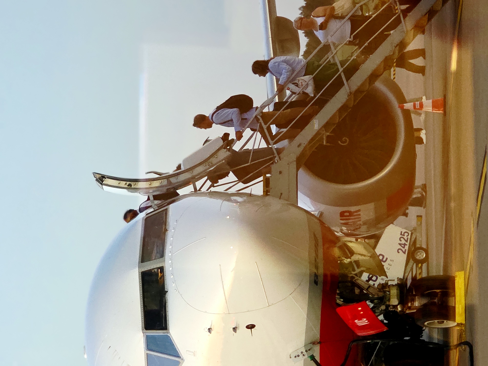 This is a close-up, angled shot of the front of a commercial airliner parked on an airport tarmac, likely at Poznań–Ławica Henryk Wieniawski Airport, Poland. Passengers are boarding the aircraft via a mobile staircase. The nose of the plane, with its cockpit windows, is prominently featured on the left side of the frame. The jet engine is visible on the right, with the boarding stairs ascending towards the aircraft's door. Several individuals, dressed in casual attire, are in various stages of boarding, carrying luggage. Some are looking towards the aircraft or the stairs, while others are focused on their ascent. The sky above is a clear, pale blue, indicating daylight, possibly late afternoon or early evening due to the warm lighting. The ground beneath the aircraft appears to be paved tarmac, with yellow markings visible. Orange traffic cones are placed on the ground near the stairs. The airline's logo and identifying text are partially visible on the aircraft and on luggage.