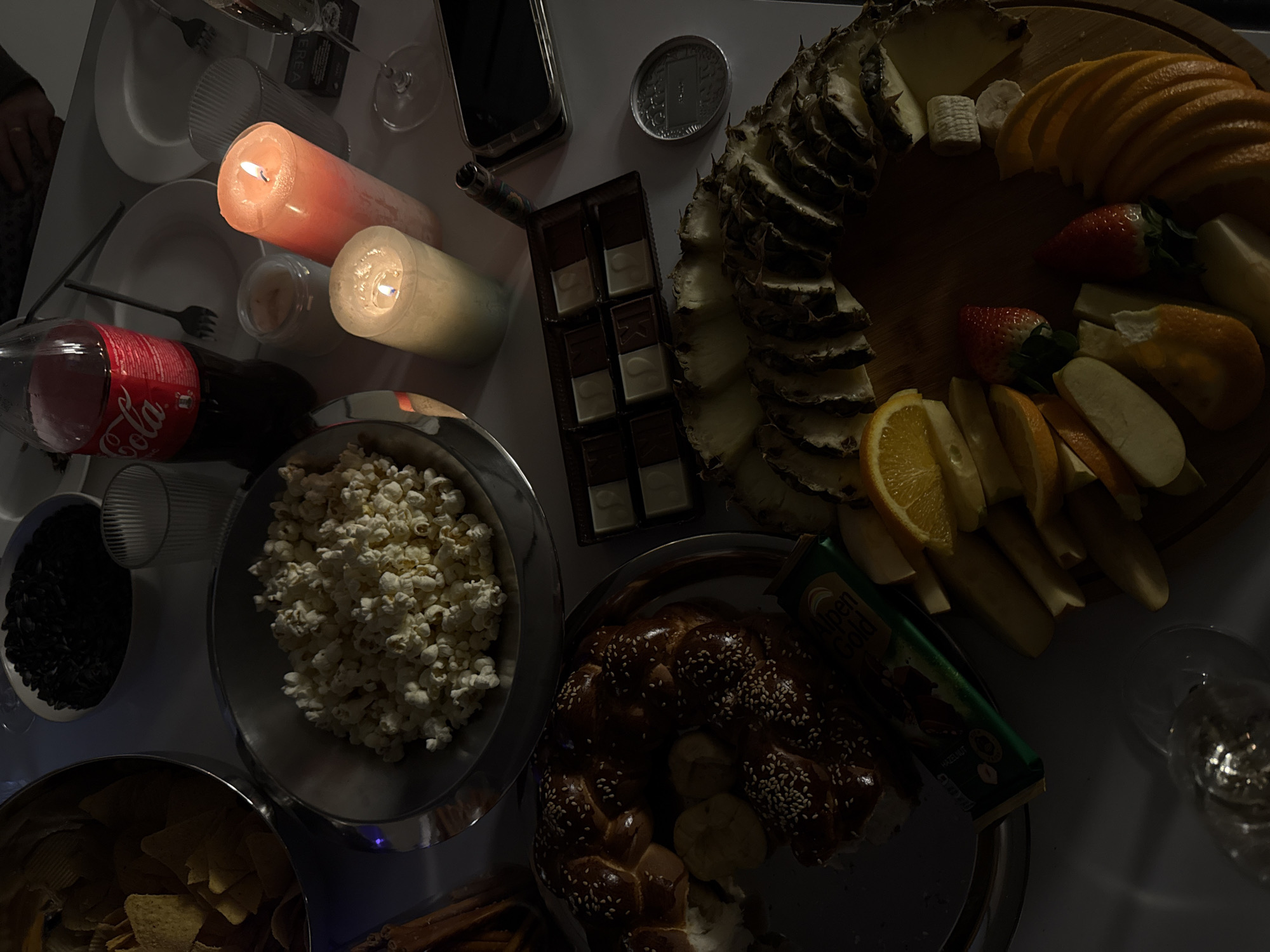 The image shows a table laden with snacks and treats, likely taken indoors under low lighting conditions with the scene illuminated by candlelight.
**Objects:**
* Two lit candles, one orange and one white, placed near a bottle of Coca-Cola and various drinking glasses.
* A mobile phone and a silver coin.
* A dark chocolate bar divided into squares.
* A wooden board featuring an assortment of fruits, including sliced pineapple, orange slices, strawberries, apple slices, and cheese.
* A plate with a braided pastry (likely challah bread).
* A candy bar next to the braided pastry.
* Bowls filled with popcorn, sunflower seeds, and potato chips/tortilla chips.
* Forks and plates are scattered on the table.
* Wine glasses are present as well.
**Subjects:**
* A hand is visible at the edge of the image.
**Actions/Interactions:**
* The scene appears to be set for a casual gathering or a snack-heavy meal. The arrangement of food indicates a sharing-style setup.
**Environment:**
* The environment appears to be indoors, possibly a dining room or living room. The dim lighting creates a cozy, intimate atmosphere.
**Location:**
* There is no information on the city of Yerevan, Armenia, in the image.