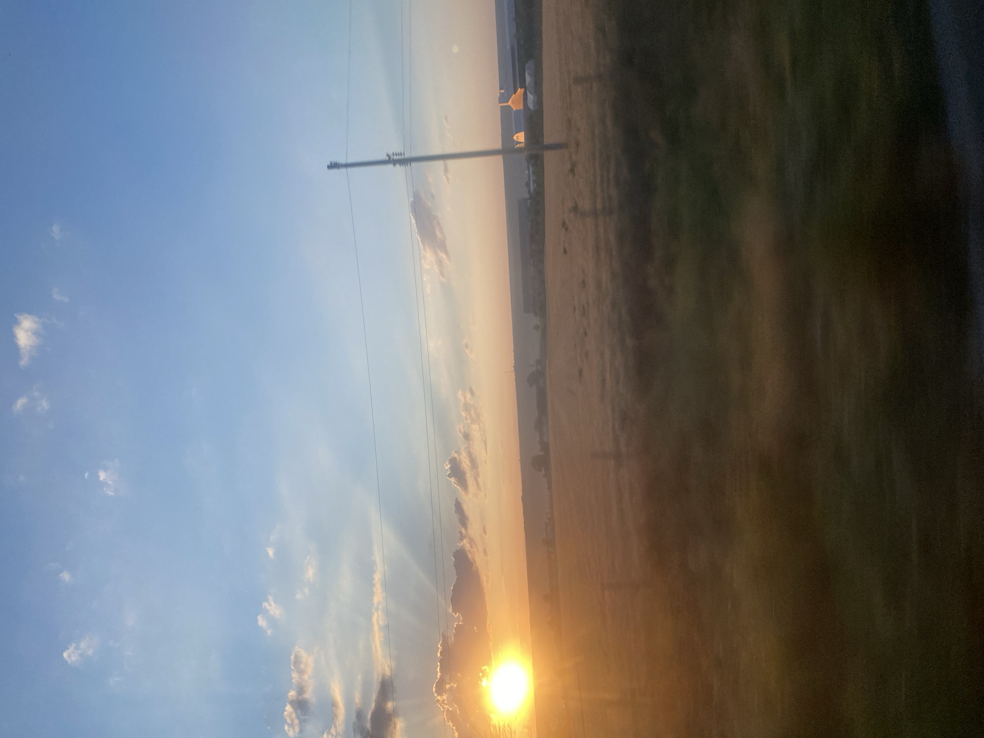 The image depicts a rural landscape at sunset, possibly in Balabanu, Moldova. The sun, a bright orb casting golden rays, is positioned low on the left horizon, partially obscured by clouds. The sky transitions from a warm orange hue near the horizon to a clear blue overhead, with scattered white clouds. In the foreground, a field of dry grass stretches towards a distant line of trees and buildings. A utility pole with power lines stands vertically in the mid-ground, slightly off-center to the right. The overall impression is one of a tranquil, late afternoon scene in the countryside.