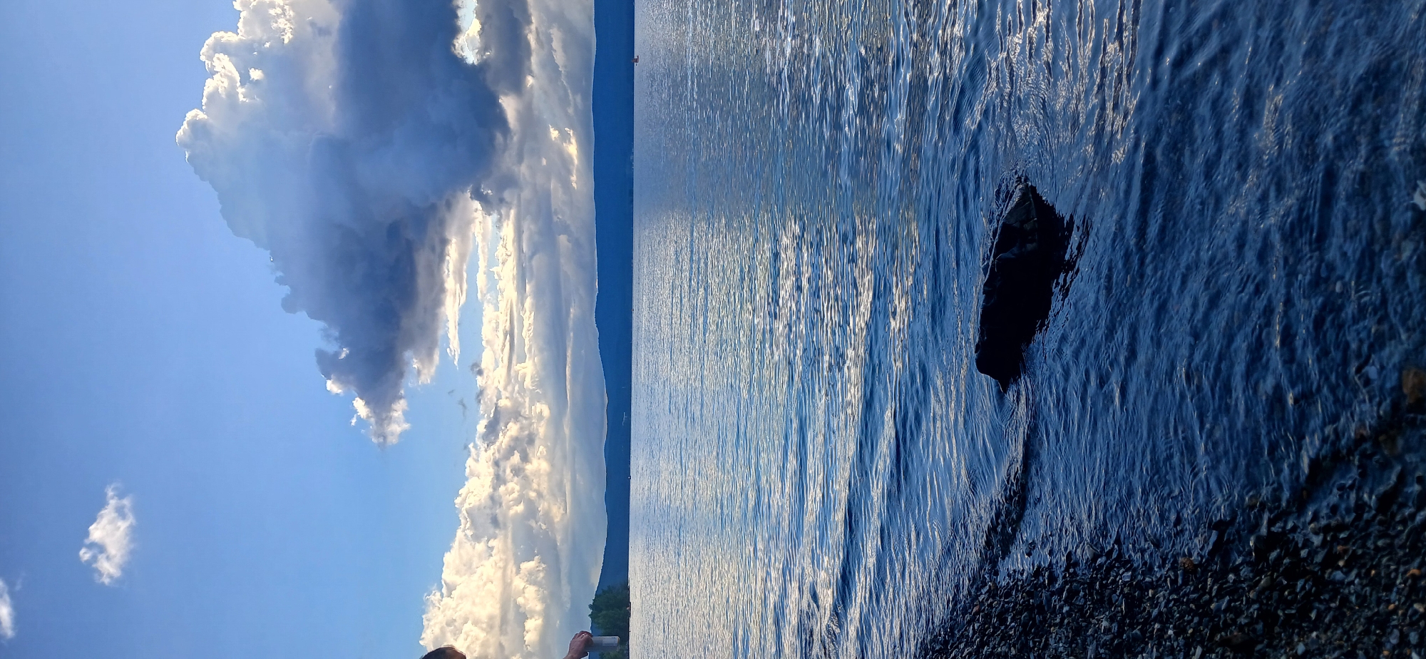 The image depicts a natural scene at the shore of Lake Turgoyak in Russia. The water is calm with gentle ripples, reflecting the sky above. A rocky shoreline with small pebbles lines the left side of the frame, leading into the water. A dark, submerged rock is visible in the foreground. In the distance, a range of mountains forms the horizon, partially obscured by atmospheric haze. The sky is a mix of blue and dramatic clouds, with some areas brightly lit by sunlight. On the far left, a person is partially visible, holding what appears to be a can. A small red object, possibly a boat or buoy, is present on the water in the distance. The overall impression is one of a tranquil, natural landscape under a dynamic sky.