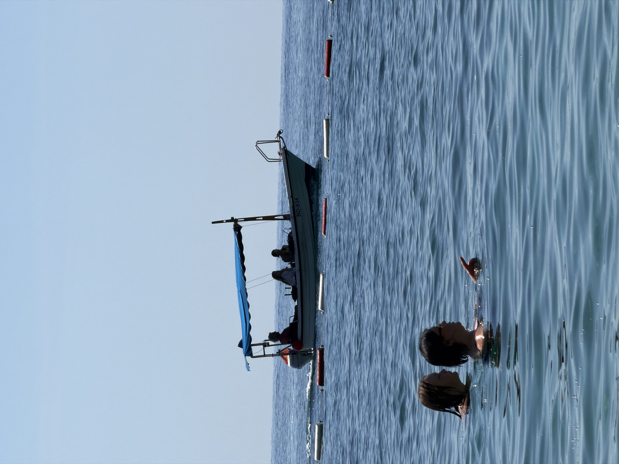 The image depicts an open water scene, likely off the coast of Budva, Montenegro, under a clear, light blue sky with no visible clouds.
In the foreground, two individuals, appearing female, are partially submerged in the calm, blue-green water. The individual on the left has light-colored, wet hair and faces right in profile. The individual on the right has shorter, dark hair, wears sunglasses, and is pointing forward and slightly right with their right index finger, facing left in profile. They appear to be interacting.
In the mid-ground, a white motorboat with a blue canopy and a red accent stripe is moving from left to right, leaving a distinct wake behind it. The boat displays the registration "BD-934" on its bow, indicating its origin is likely Budva. An outboard motor, identifiable as a Honda, is mounted at the stern. At least three occupants are visible as dark silhouettes under the blue canopy, with one person appearing to operate the boat from the left side. A line of cylindrical buoys, alternating between white and red, stretches across the water, positioned between the swimmers and the boat, marking a designated area. The horizon line between the sea and the sky is clearly visible in the background.