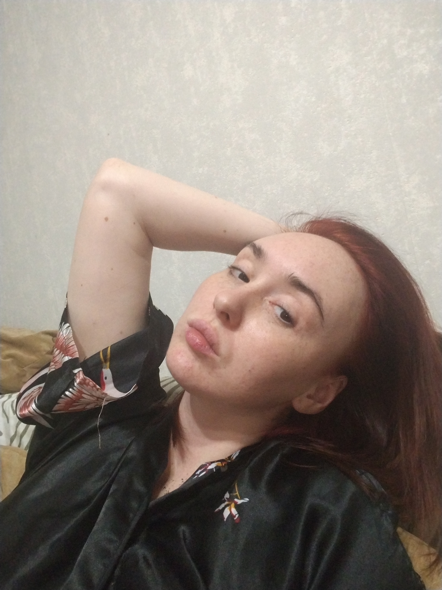 A pale-skinned woman with red hair, freckles, and prominent eyebrows is reclining on what appears to be a bed or soft surface. She is wearing a black satin robe with a floral and crane design. Her left arm is raised above her head, and she is looking at the camera with slightly pursed lips. The background is a plain, light-colored wall. The photo appears to be taken indoors. There is no verifiable evidence that the image was taken in Izhevsk, Russia.