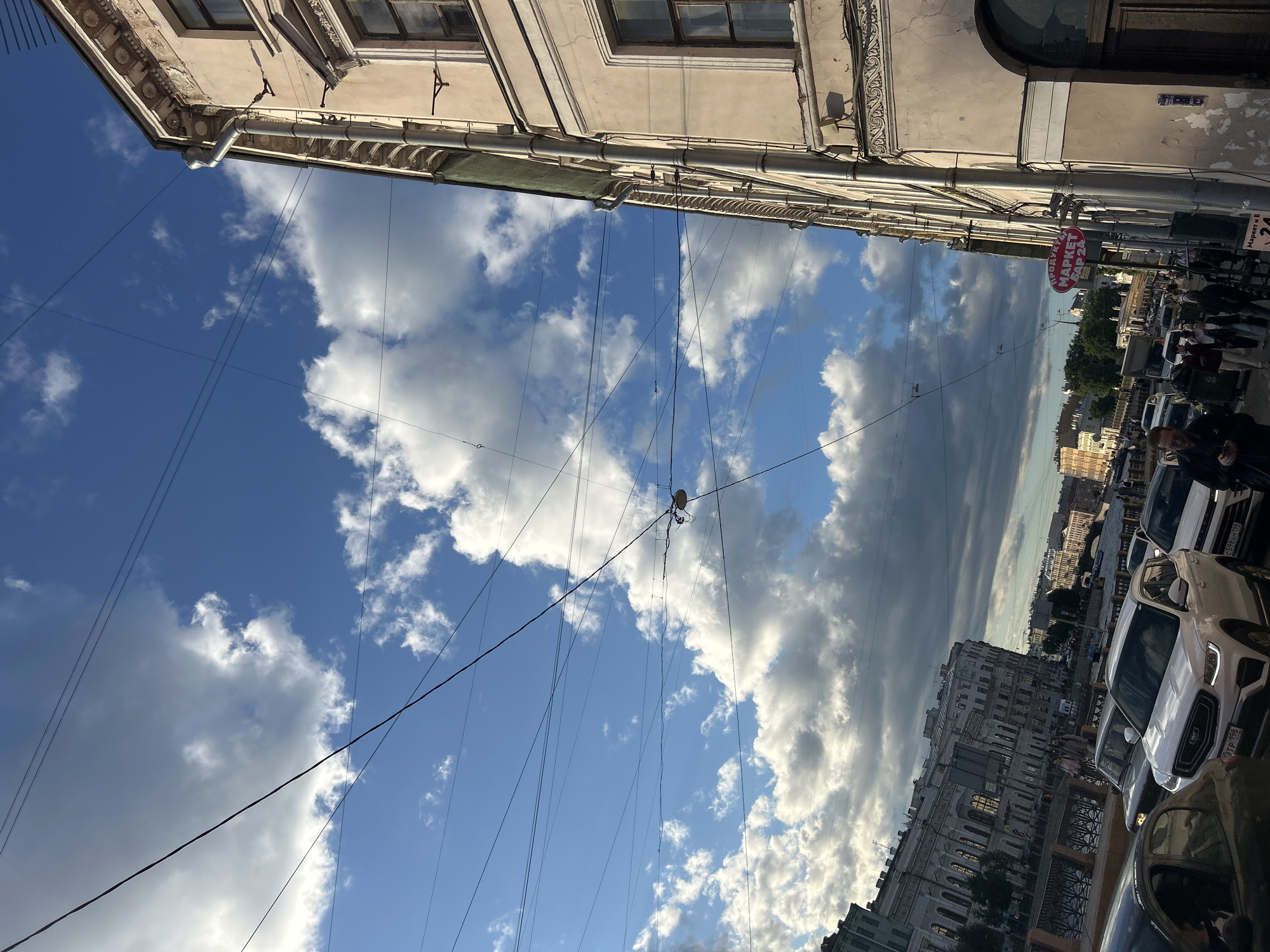 The image captures a street scene in an urban environment with a prominent view of the sky, characterized by scattered white clouds against a bright blue backdrop. Overhead, a complex network of telephone or electrical wires crisscrosses the frame.
On the right side of the image, a building with an ornate facade stands tall, featuring detailed architectural elements such as cornices and window frames. A metal downspout runs vertically along its exterior. Below, a sign with Cyrillic text, "ПРОДУКТЫ МАРКЕТ", is visible, indicating a local market or shop.
The left side of the image reveals a canal or waterway with a decorative fence along its edge. Across the water, a grand, multi-story building with numerous windows lines the embankment. Further in the background, more urban structures and trees can be seen, suggesting a city with a rich architectural history.
The street level shows moderate traffic, with several cars, including a white SUV, a dark sedan, and other vehicles, navigating the roadway. Pedestrians are also present, with individuals walking on the sidewalk. The overall atmosphere suggests a lively city street during daylight hours, possibly in the late afternoon given the lighting.