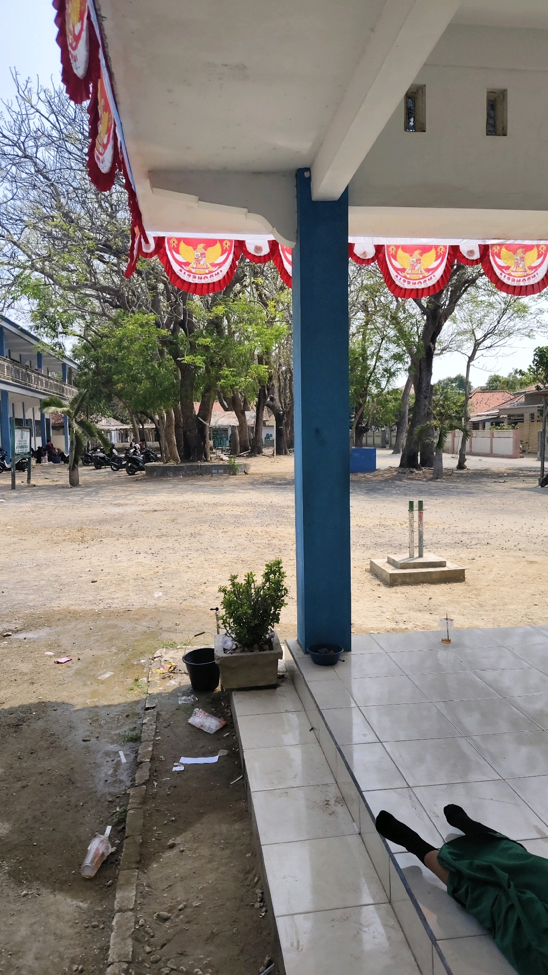 The image depicts an outdoor scene in Pangpajung, Indonesia, likely a school or public building courtyard. In the foreground, a person's legs wearing dark socks and green trousers are visible, lying on a tiled floor. To the left of the tiled area, a dirt and gravel ground is visible with scattered debris including plastic bags and a disposable cup. A potted plant sits on a raised tiled platform next to a blue pillar. Beyond this, a sandy courtyard stretches out, lined with trees and a building with multiple stories on the left. Several motorcycles are parked in the background, near the building. Decorative red and white banners, featuring the Indonesian coat of arms, are hung from the overhang of the building's roof. The sky is clear and bright, indicating a sunny day. The overall environment suggests a dry, possibly tropical climate.