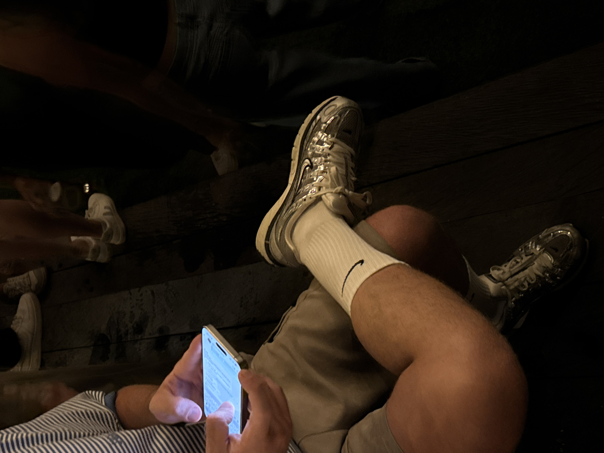 The scene depicts a person sitting on a wooden surface, possibly a deck or floor, while using a mobile phone. The person is wearing shorts and white Nike socks with silver sneakers. The phone screen is illuminated, suggesting it is in use. In the background, there are partial views of other people's legs and footwear, including at least two pairs of white shoes, positioned on the same wooden surface. The lighting is dim, casting shadows and obscuring details. There is no explicit information within the image itself confirming the location as North Kuta, Indonesia.