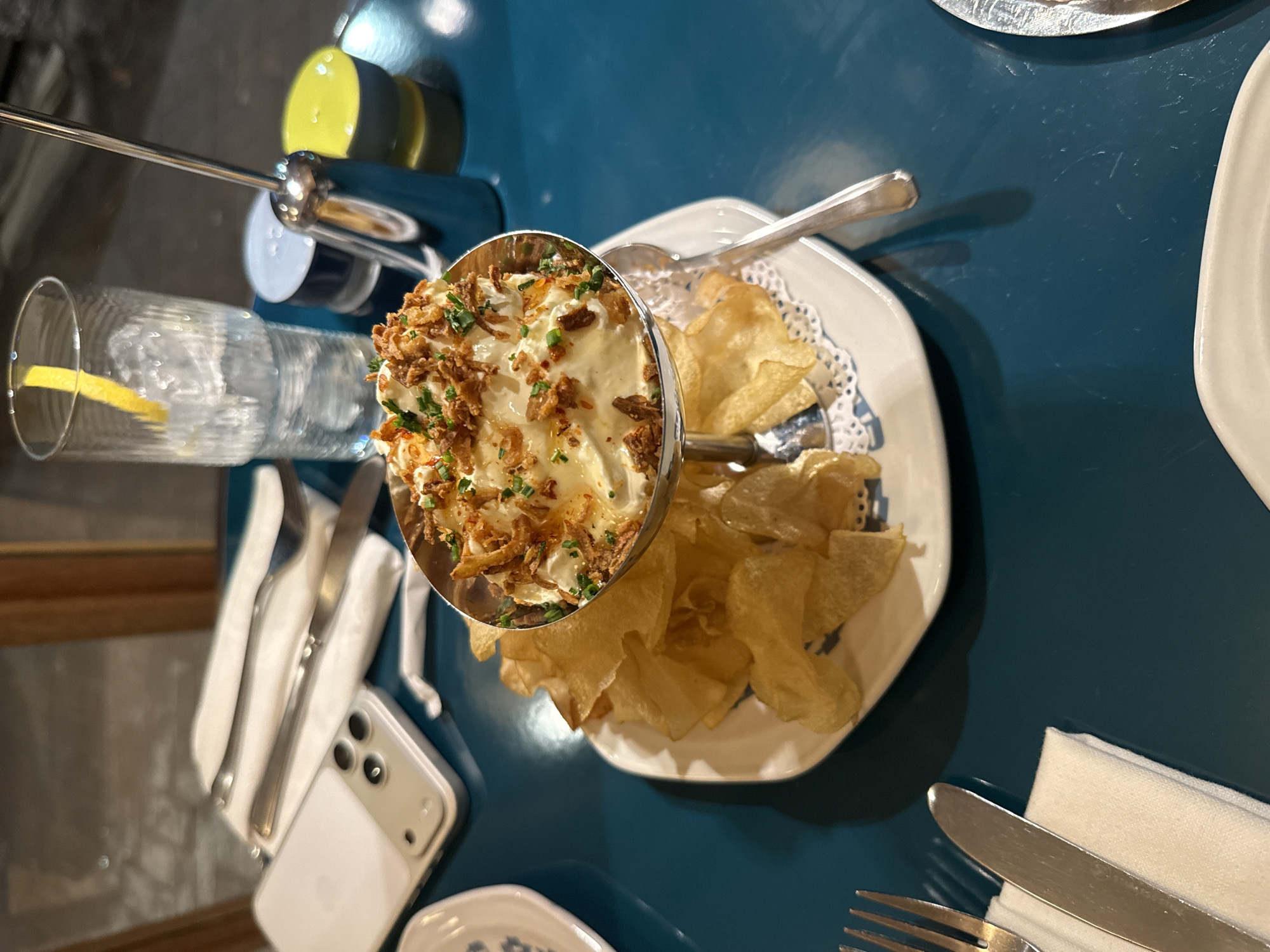 An overhead shot captures a dining scene indoors, likely at a restaurant or bar, with a prominent blue tabletop. The central focus is a small silver bowl filled with a creamy dip, generously topped with crispy fried onions and chopped chives. Surrounding the bowl, on a white, octagonal plate with a decorative doily pattern, is a scattered array of golden potato chips, ready for dipping.
To the left of the dip, a clear glass contains water with ice and a lemon wedge. Further left, partially out of frame, a white iPhone with a light-colored case rests on the table next to a folded white napkin and silverware (a fork and a knife). Another set of cutlery and a napkin are visible on the far right side of the table.
In the upper left background, some shiny metal objects are visible, including what appears to be a dispenser or container with yellow accents, reflecting the ambient light. The lighting suggests an indoor setting, possibly evening, with artificial lights creating reflections on the table and metallic surfaces. The overall impression is a casual but stylish dining experience. No discernible text is visible on any objects. While no specific visual cues confirm it, the scene is set in Cape Town, South Africa.