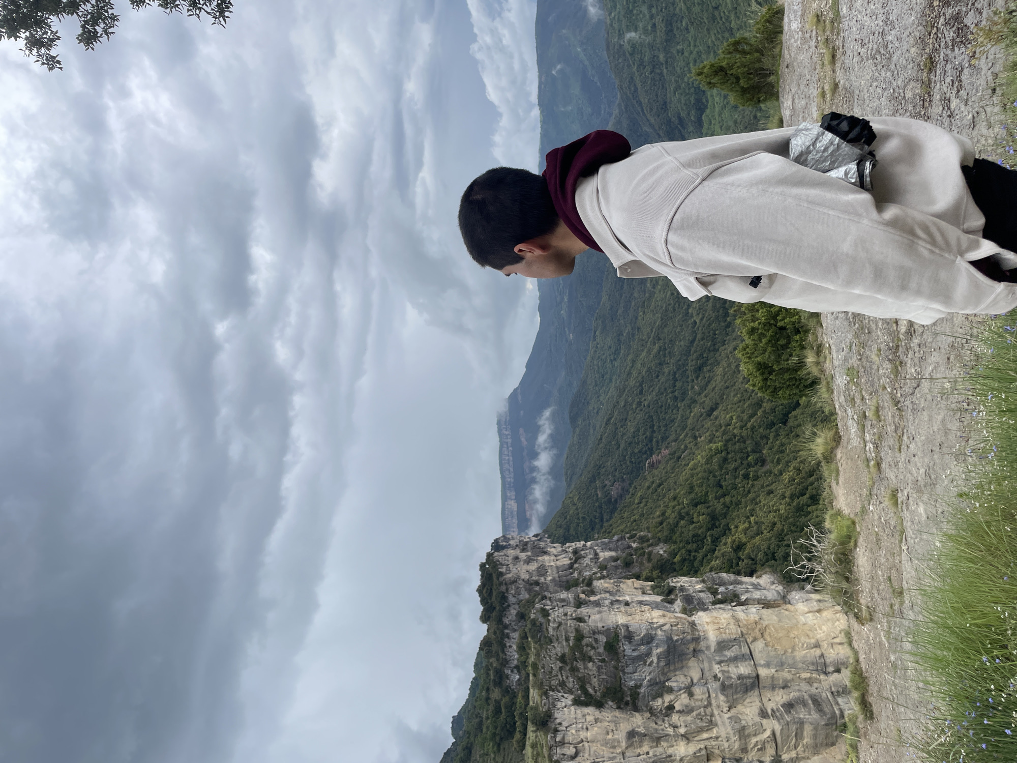 A man stands on a rocky precipice, gazing over a dramatic landscape of forested mountains and a sheer cliff face, beneath a cloudy sky. He wears a light-colored jacket with a burgundy hood. The location appears to be near Rupit i Pruit, Spain.