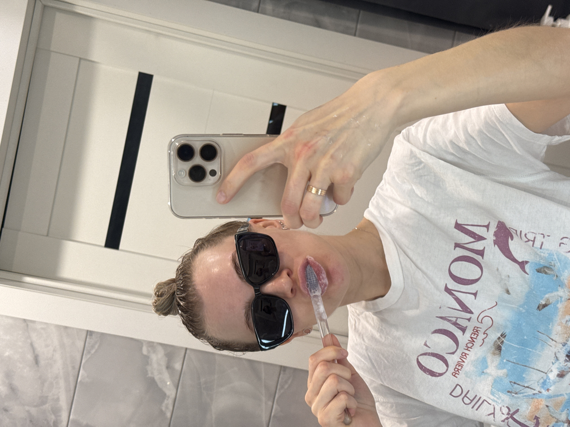 A woman is in a bathroom taking a selfie in a mirror. She is wearing sunglasses and a white t-shirt with "MONACO" printed on it in a mirrored orientation. She is holding a toothbrush with a pink substance on it to her lips. In her other hand, she is holding a smartphone with three visible camera lenses. She has fair skin, and her hair is pulled back in a bun. Her left hand has a gold ring on her ring finger. The bathroom has white walls and white cabinets. There is no visible evidence to indicate this photo was taken in Izhevsk, Russia.