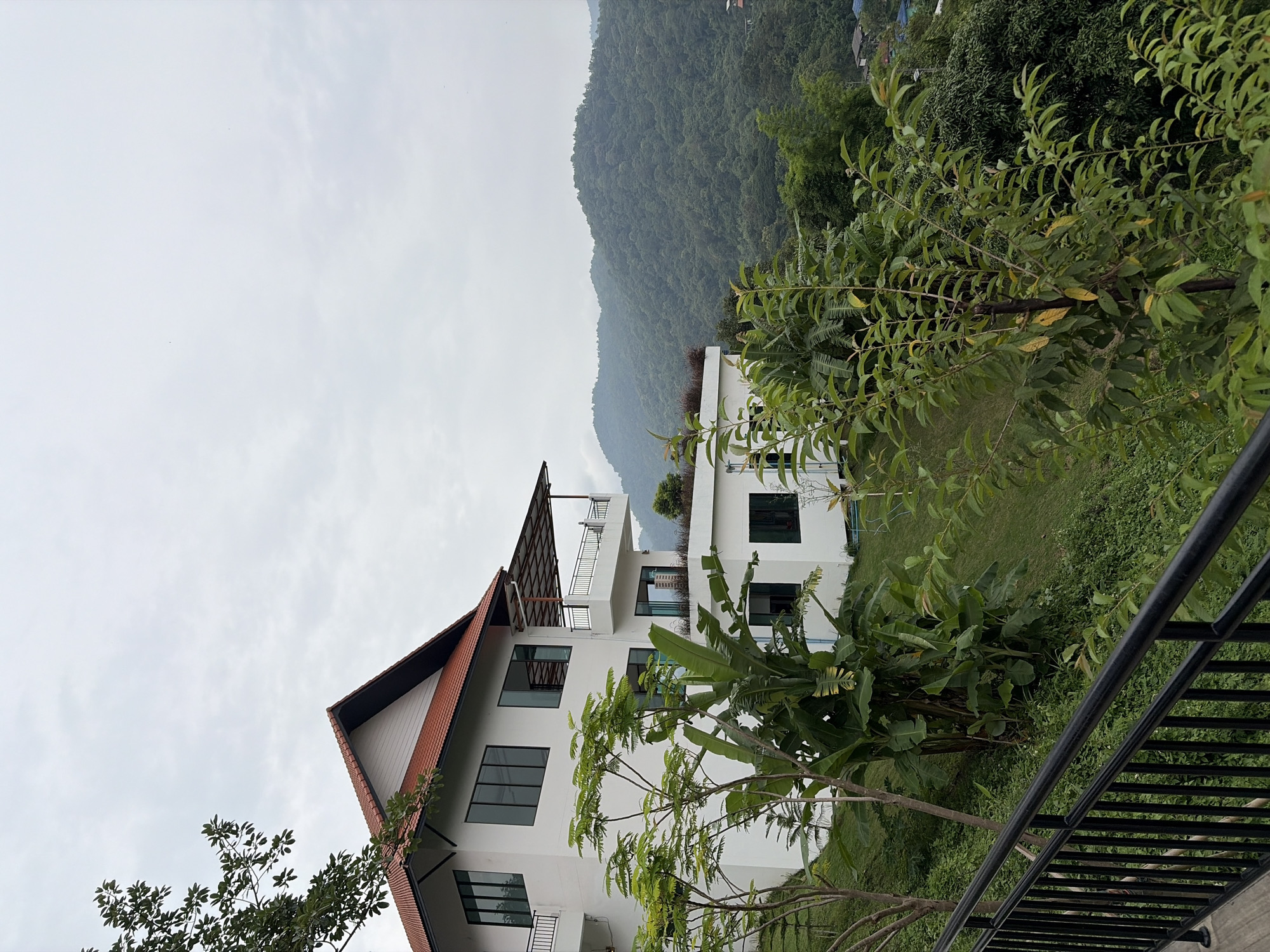 A white multi-story building with a red tile roof is situated on a hillside overlooking a lush, tree-covered mountain range. The building has several balconies with black railings, large windows, and is surrounded by abundant green vegetation, including trees and banana plants. A black metal railing is visible in the foreground, leading up to a wooden walkway. The sky is overcast and gray. The location is identified as Ban Kao Dua, Thailand. There are no people visible in the image.