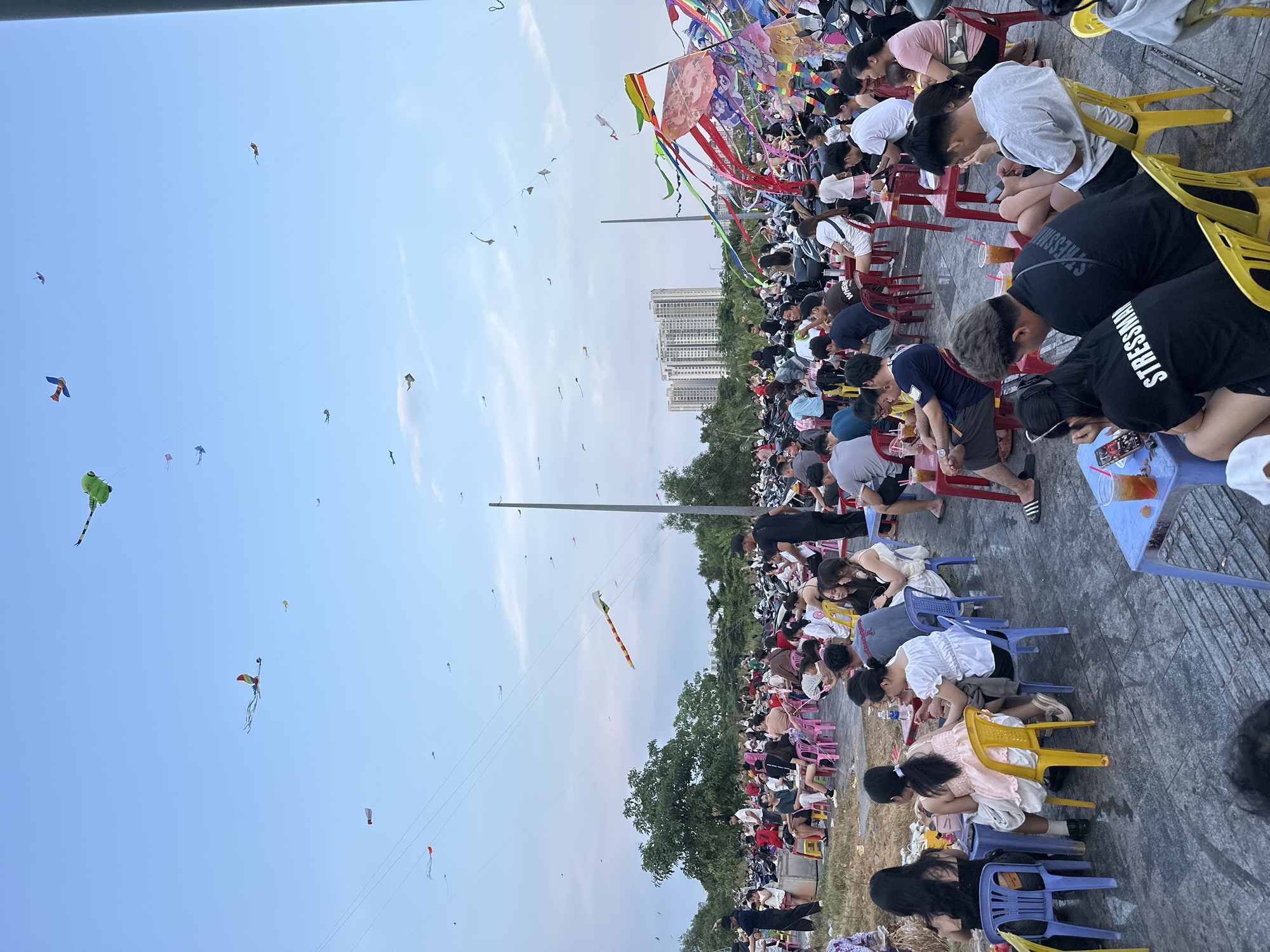 The image captures a kite flying festival held in Thủ Đức, Vietnam. A large crowd of people, seated on colorful plastic chairs, stretches across the scene. Many are engrossed with their phones or drinks on small tables. Above them, a multitude of kites of varying colors and designs populate the sky. A line of trees provides a backdrop with a few modern buildings visible in the distance. The sky is a gradient of pale blue.