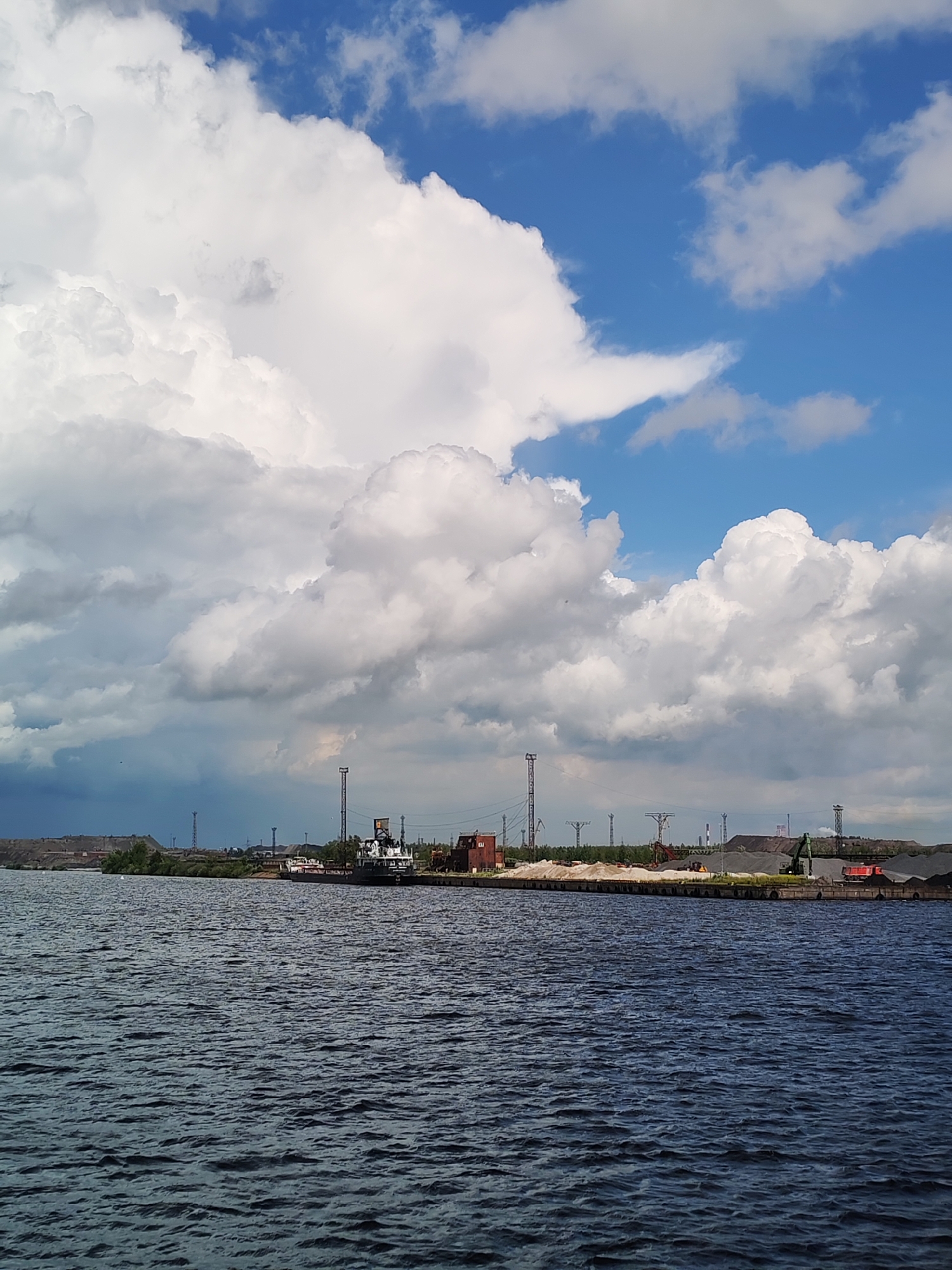 The image shows a wide body of dark blue water in the foreground, with visible ripples on its surface. In the background, a distant industrial waterfront is visible under a dramatic sky filled with large, puffy white clouds and patches of blue. The waterfront features several structures, including industrial cranes, tall metal towers, piles of gravel or coal, and buildings. A large white and dark-colored vessel is docked along the waterfront, with another smaller boat nearby. The overall scene suggests an industrial port or facility located on a river or lake. This location is likely within the city of Cherepovets, Russia, known for its significant industrial complexes.