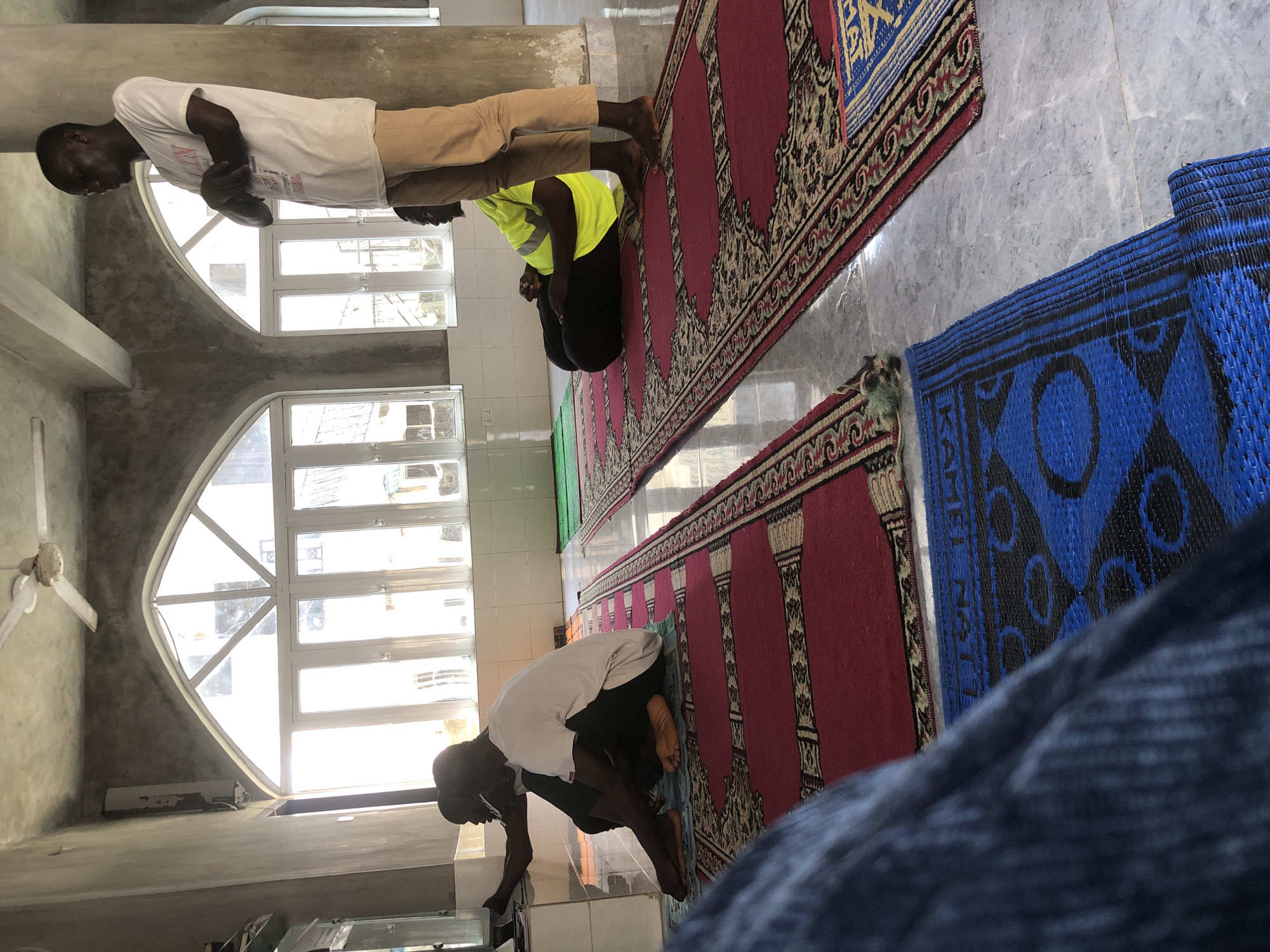 The image shows the interior of a room, possibly a mosque, with a high ceiling and large arched windows that let in bright sunlight. The walls are bare concrete, and a ceiling fan is visible. Several people are present. In the foreground, a person's legs are covered by dark blue fabric, suggesting they are sitting or lying down. To the right of the foreground, three men are visible. One man stands with his arms crossed, wearing a white t-shirt and light brown pants. Another man is seated on the floor, facing away from the camera, wearing a yellow high-visibility vest and dark pants. A third man is seated next to him, facing towards the left, wearing a white t-shirt and dark pants. The floor is covered with patterned rugs in red and blue. The overall scene suggests a gathering or prayer session.