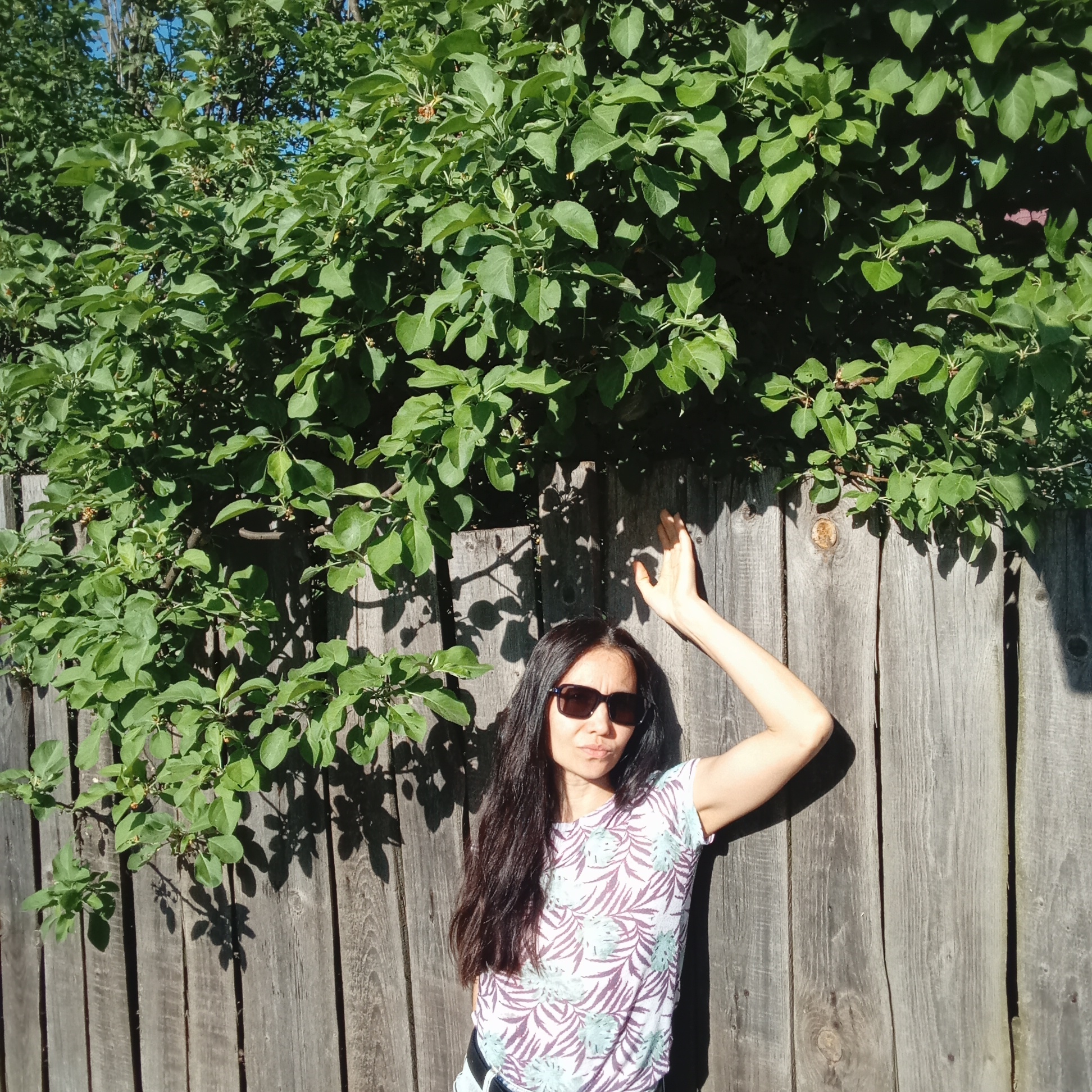 A woman with long dark hair and sunglasses is standing against a wooden fence in front of a leafy green tree. She is wearing a white t-shirt with a palm leaf pattern. Her right arm is raised and resting against the fence. The location is Буренка, Russia.