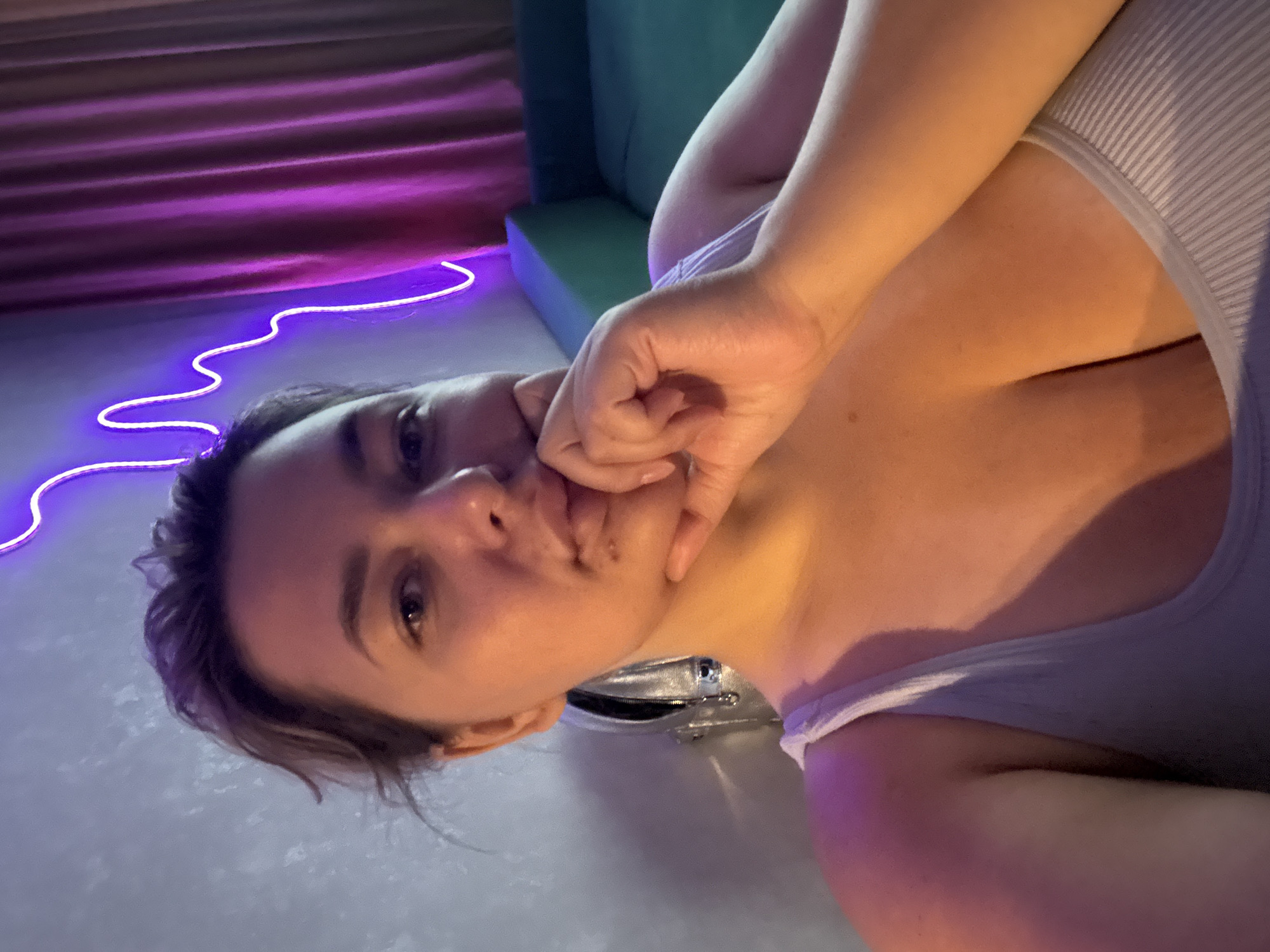 A young woman is lying down, with her head tilted and her hand resting on her face. She is wearing a light-colored tank top. The background features a purple curtain and a neon light. The setting appears to be indoors, possibly a bedroom or a lounge area, with a hint of a green couch in the background. The lighting is dim, with a mix of ambient light and the vibrant glow of the neon. The overall mood is relaxed and intimate, with the woman's pose and expression suggesting a moment of quiet contemplation or playfulness. The image is oriented sideways, adding to its unconventional and artistic feel. There is no visible text in the image.