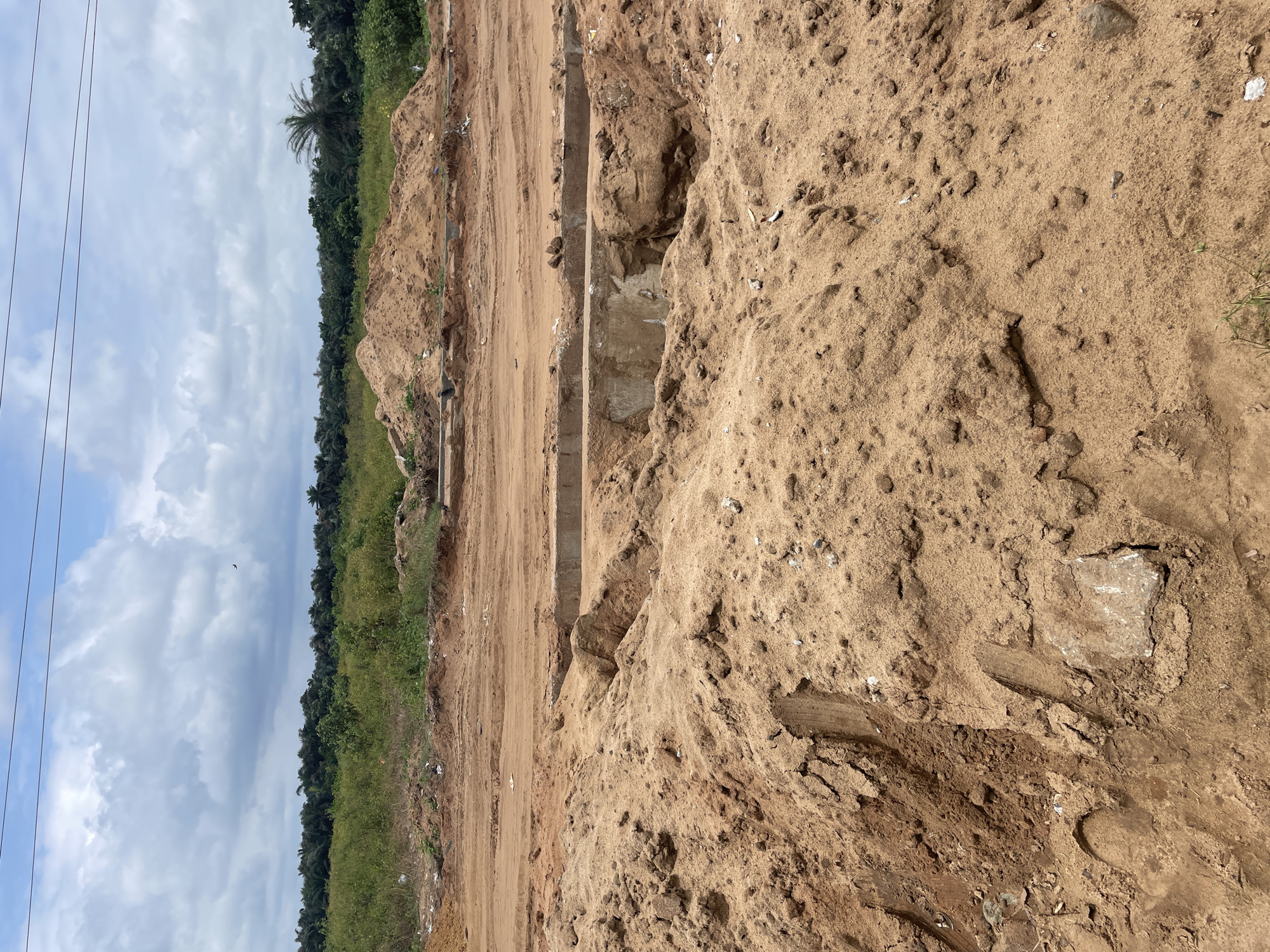 The image depicts a construction site with large mounds of sand and soil in the foreground. In the midground, there is a partially constructed concrete structure, possibly a drainage channel or foundation, with exposed rebar. The background features a dense line of trees and vegetation, indicative of a tropical environment. The sky above is partly cloudy with power lines stretching across the top of the frame. The scene is located in Iyafin, Nigeria, as indicated by the context provided. There are no people or animals visible in the image. The overall impression is one of ongoing civil engineering work in a rural or semi-rural setting.