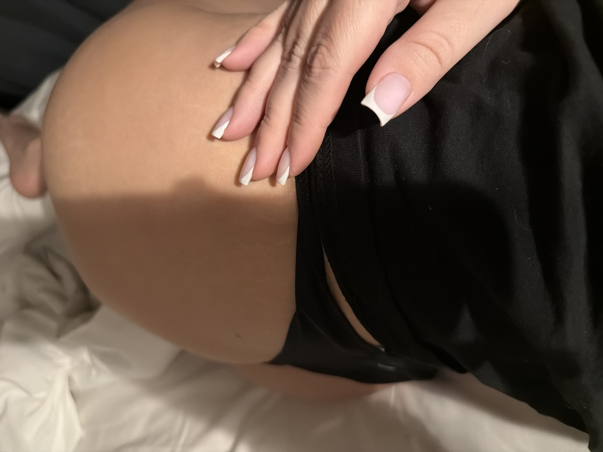 A close-up shot depicts a person lying on a white bed, partially covered by black clothing. The person's buttocks are visible, and a hand rests on the skin near the edge of the clothing. The hand has manicured nails with a white tip design. The background is out of focus, but there is a hint of what appears to be a dark curtain. The location in Moscow, Russia is not discernible.