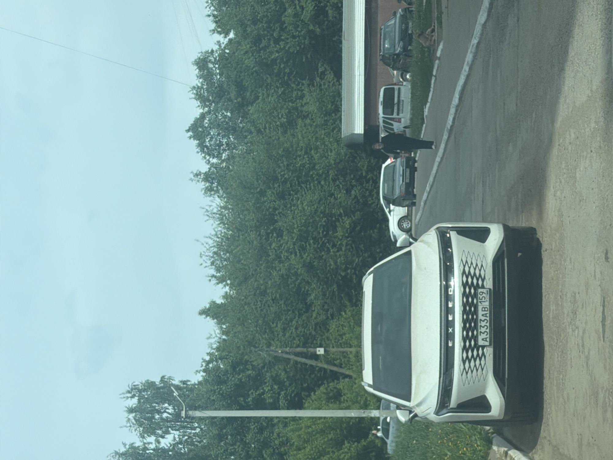 The image shows a street in Chaikovsky, Russia. A white Exeed TXL car with license plate "A333AB 159" is parked on the right side of the road. Further down the road, parked along the same side, are at least 3 other vehicles of various sizes including one that is dark grey, another that is white. An individual in dark clothing is walking on the sidewalk towards the darker-colored vehicles. A line of trees is on the left. The sky is overcast. A utility pole is also visible on the left.