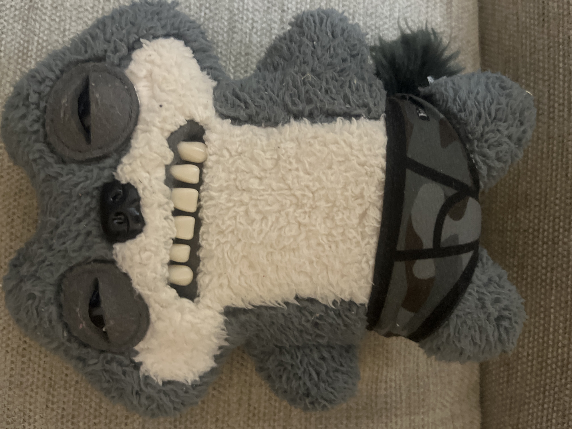 This is a close-up, eye-level, medium shot of a plush toy that appears to be a raccoon or similar creature. The toy is predominantly grey and white, with a fluffy texture. It has large, round, grey eye sockets with black button eyes that have an open slit, giving it a sleepy or exhausted appearance. The toy has a prominent snout and a wide mouth with numerous white, square teeth, suggesting a goofy or surprised expression. It is wearing a pair of grey camouflage-patterned underwear. The toy is positioned on a textured, beige fabric surface, likely a sofa or carpet. The lighting is neutral, and there are no visible shadows suggesting a specific time of day. The image does not depict any specific location in Aktau, Kazakhstan, or any identifiable human presence.