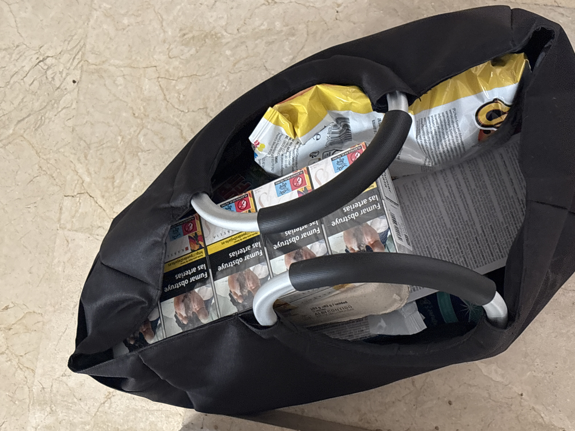 The image shows an open black bag containing several items, including a bag of snack chips and multiple packs of cigarettes. The cigarettes are stacked vertically and some of the warning labels are visible. The bag is resting on a tiled floor. The overall context of the scene is domestic, and there is no indication of it being in the city of Orihuela, Spain.