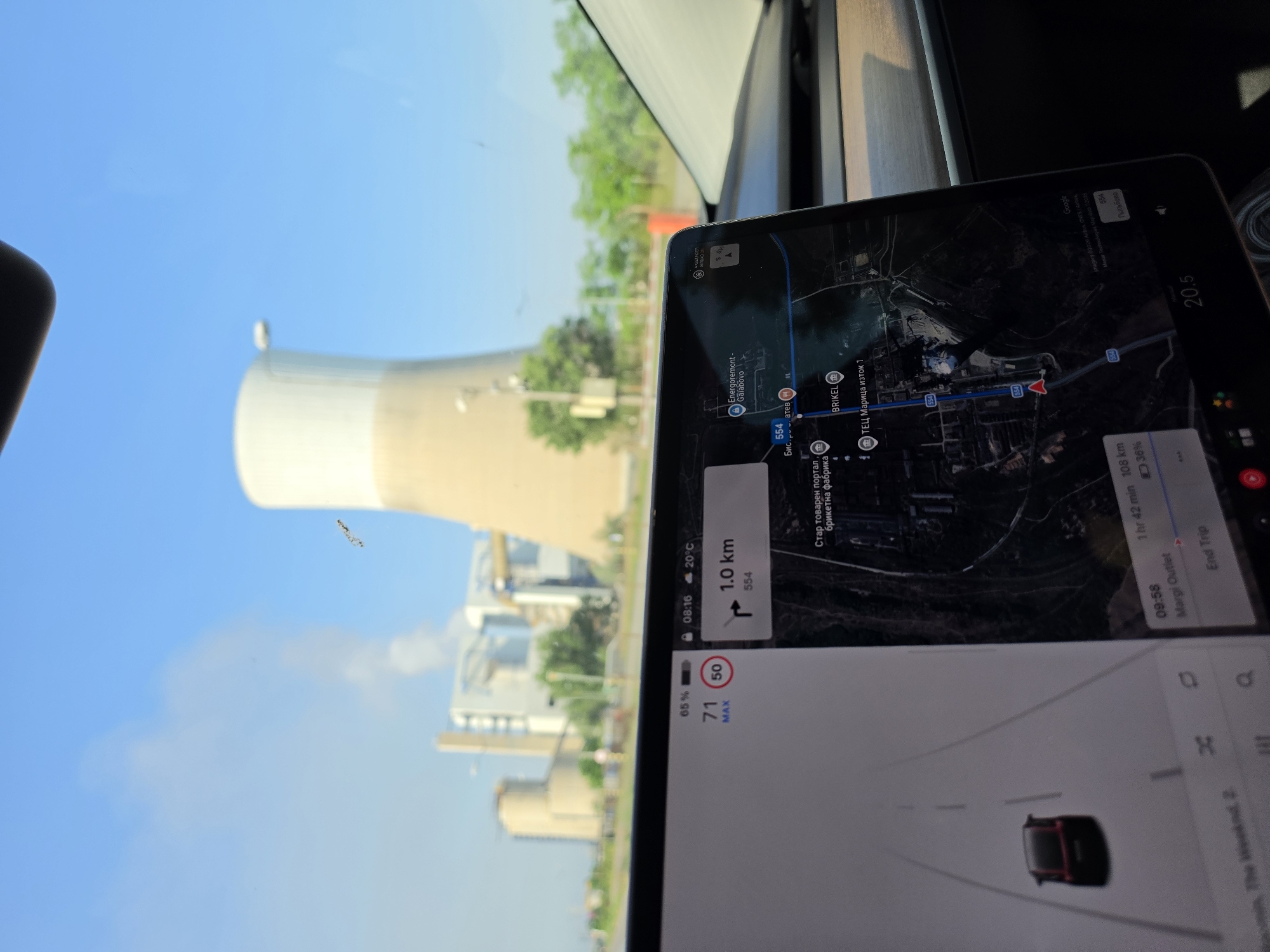 The image depicts a scene observed from inside a vehicle, looking towards an industrial power plant under a clear blue sky. A large digital display screen, characteristic of a Tesla vehicle, occupies the lower foreground.
In the background, a substantial industrial complex is visible. A prominent, light-colored cooling tower, with a white upper section and a beige base, dominates the center-right. To its left, several other industrial buildings, including a large, darker structure and a cylindrical silo, are present. Plumes of white vapor or smoke ascend into the sky from chimneys situated behind these structures. Green trees and foliage are visible along the horizon line to the right of the power plant.
The vehicle's central touchscreen displays navigational and operational information. The navigation map shows a route with a pending left turn in 1.0 km on road 554. Key locations indicated on the map include "Energoremont - Galabovo," "БРИКЕЛ" (Brikel), and "ТЕЦ Марица изток 1" (TPP Maritsa Iztok 1), confirming the geographic location as the vicinity of Galabovo, Bulgaria. The display indicates a current speed of 71 km/h, exceeding the posted speed limit of 50 km/h. Other readouts include a battery charge of 65%, the time 08:16, and an ambient temperature of 20°C. The estimated journey duration is 1 hour 42 minutes covering 108 km, with "Margi Outlet" as the destination. A stylized representation of the vehicle is shown on the left side of the screen, moving along a digitized road. The vehicle's dashboard is visible below the screen, and a portion of the windshield shows reflections of the sky. A small, dark, elongated object is present on the windshield, positioned against the blue sky to the right of the main cooling tower. The sky is bright blue with some diffused white vapor from the power plant.