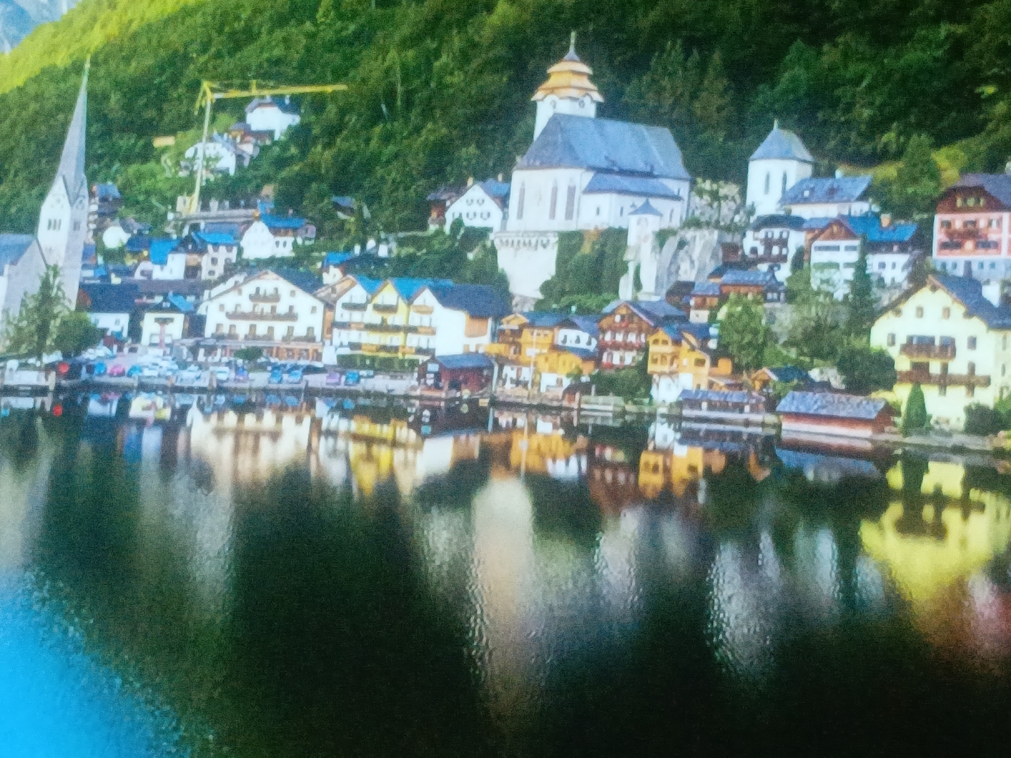 The image shows the town of Hallstatt, Austria, reflected in the Hallstätter See lake. The town is nestled against a green hillside, with buildings of various colors and styles clustered together. Prominent features include a church with a tall spire, a larger building with a domed tower, and numerous houses with balconies and visible parking. A yellow construction crane is visible in the background. The lake surface is mostly dark, but reflects the buildings and their colors.