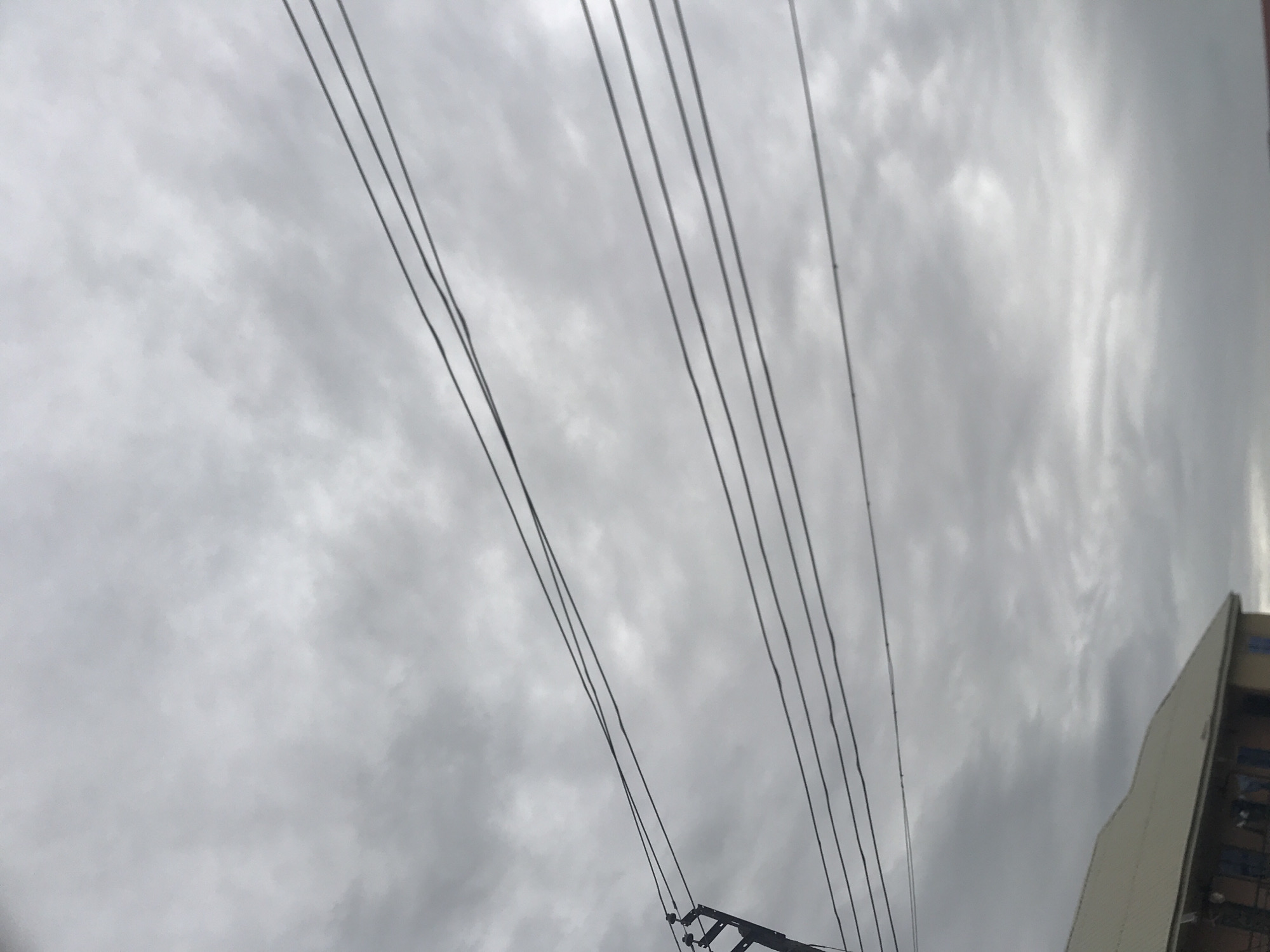 The image shows an overcast sky with multiple power lines stretching across the frame. A portion of a building with a light-colored, sloped roof is visible in the lower left corner. The setting appears to be an urban or semi-urban environment. The specific location is Rafin Par, Nigeria. The overall atmosphere is muted due to the cloudy sky. No people are visible in the image.