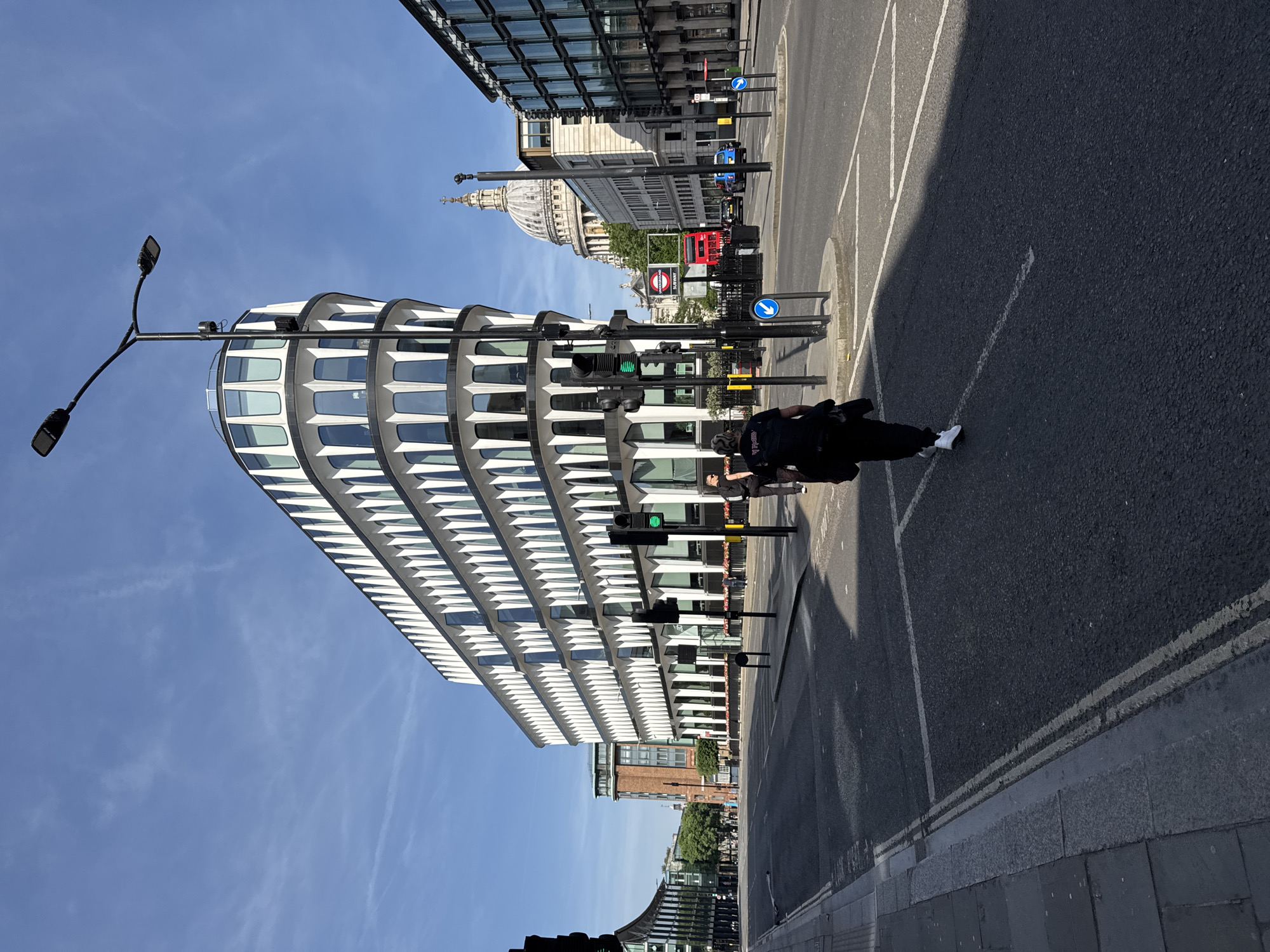 This image depicts a street scene in London, United Kingdom, featuring a modern building with a white, tiered facade on the left side. A street runs vertically through the frame with a person wearing dark clothing and white sneakers walking on the right side. Traffic lights are visible in the center, indicating the street is active. In the background, St. Paul's Cathedral can be seen, along with a red double-decker bus and other buildings. The sky is blue with few clouds. A lamppost is visible on the left, extending outwards.