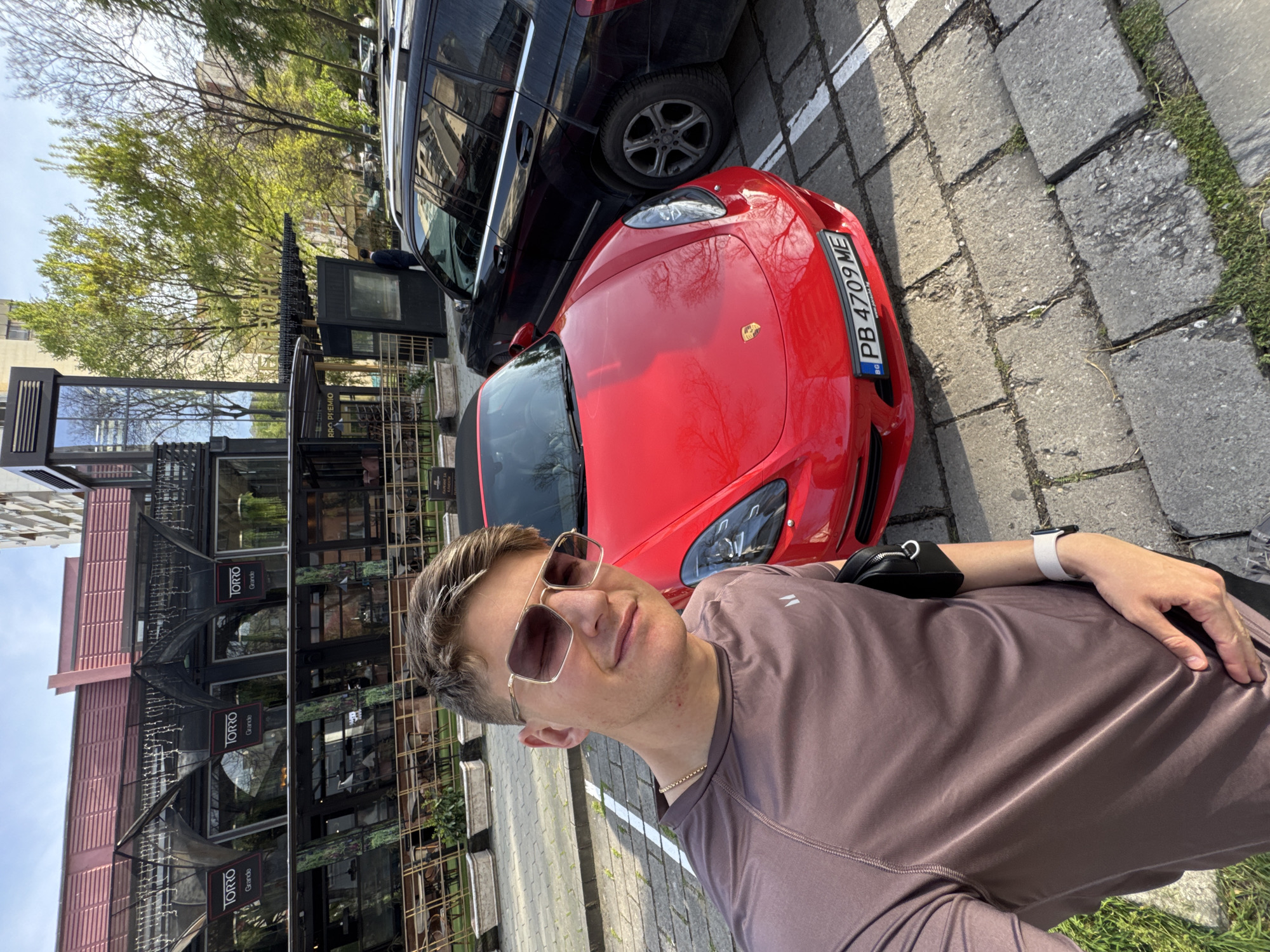 A man with short brown hair, sunglasses, and a brown t-shirt stands outdoors, likely taking a selfie. He is positioned near a red Porsche parked next to a black SUV on a paved road. The car's license plate reads "PB 4709 ME." The background includes a modern building with the name "TORRO Grande" displayed on multiple signs, suggesting a restaurant or commercial establishment. The presence of paving stones and greenery along the road indicates an urban environment. Based on the "PB" license plate prefix, this location is in Plovdiv, Bulgaria.