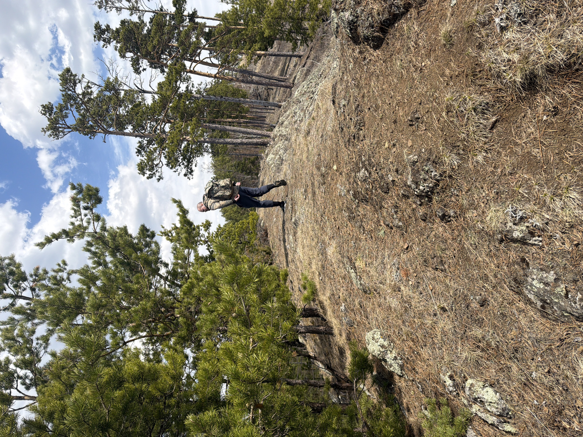 A man with a backpack stands on a rocky, pine needle-covered area, which appears to be a cliff or high ground, judging by the steep slope. He is wearing dark clothing and a camouflage backpack. The background includes pine trees and a blue sky with clouds. The location is in the city of Невон, Russia.