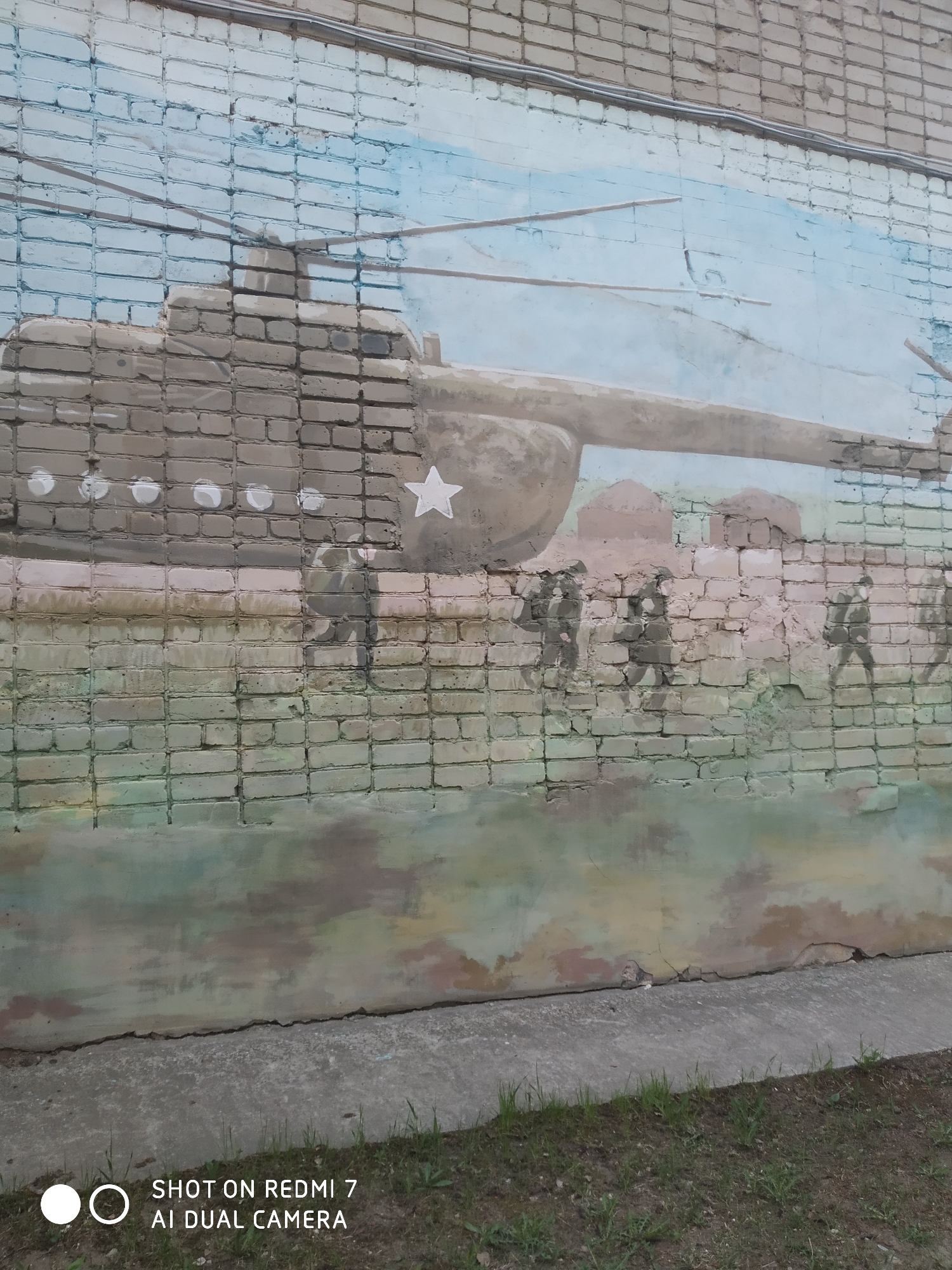 A mural is painted on a brick wall in Chaikovsky, Russia. The mural depicts a tank with a white star on its side, followed by a group of soldiers in silhouette walking forward. Above the tank, a helicopter is rendered in the sky. The bottom of the wall is covered in a mottled green and brown paint, suggesting ground or vegetation. At the base of the wall is a strip of concrete, followed by a patch of grass.