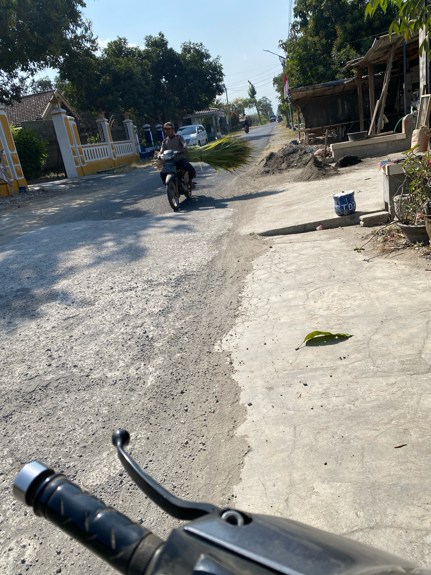 A man rides a motorcycle on a paved road in Gurah, Indonesia. He is wearing a brown cap and a gray shirt and is carrying a large bundle of green leaves on the back of his motorcycle. The road is lined with houses and trees. In the foreground, the handlebars of another motorcycle are visible, suggesting the photo was taken from a moving vehicle. A pile of sand and a blue bucket are on the right side of the road. The sky is clear and blue.