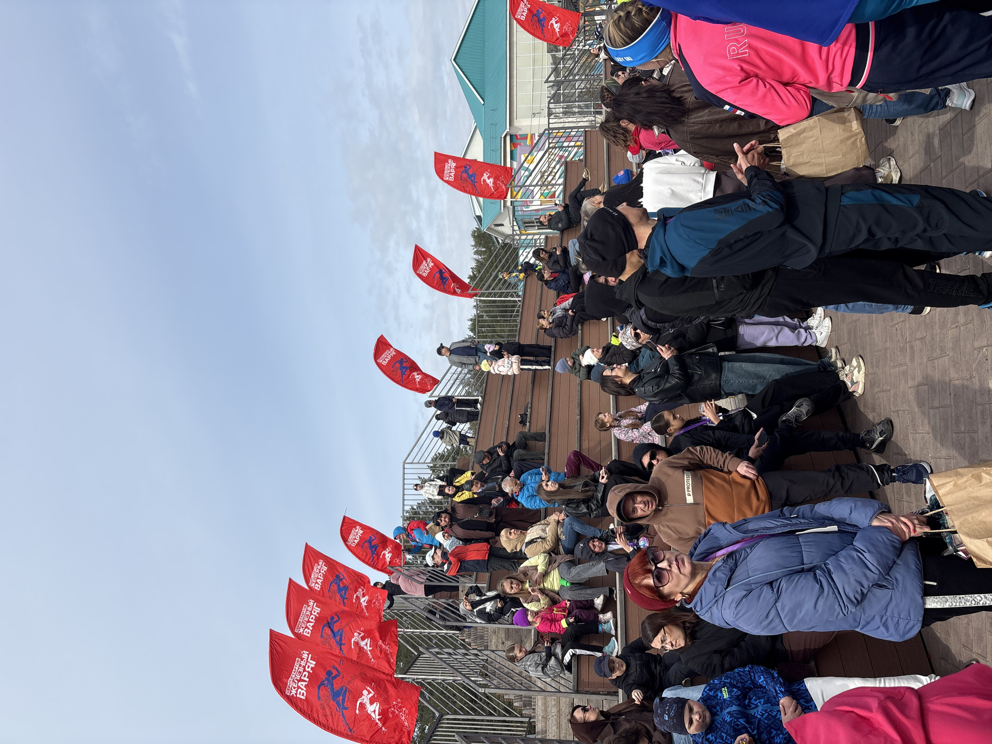 The photograph shows a large group of people sitting and lying down on tiered wooden seating, possibly bleachers or an outdoor stage, under a partly cloudy sky. Red flags with a blue silhouette of a running figure and the words "ЖЕЛЕЗНЫЙ ВАРЯГ" (Iron Viking) are positioned along the railing of the seating area. Most people are dressed in casual, cold-weather clothing like jackets, hoodies, and hats. Some appear to be resting or waiting.
The setting appears to be outdoors, near a building with a green roof. Based on the text on the flags and additional context, the location is likely in the city of Neftekamsk, Russia, where the "Iron Viking" series of races is organized.