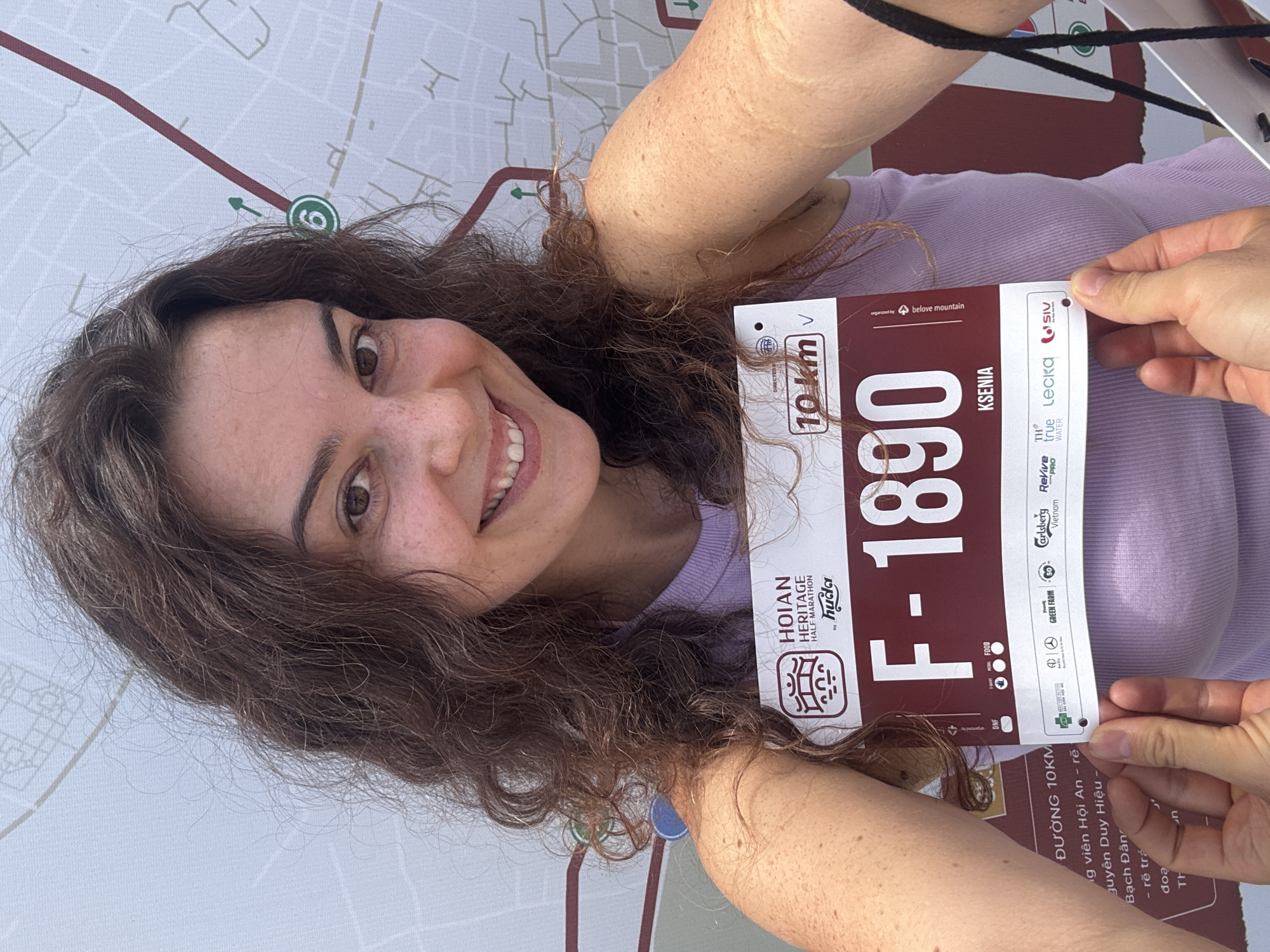 A woman with long, curly brown hair and fair skin smiles at the camera, holding a race bib. The bib is maroon and white and features the text "HOIAN HERITAGE HALF MARATHON," a race number "F-1890," and the name "KSENIA." A map of the city of Hoi An, Vietnam, is visible in the background. The woman is wearing a light purple tank top. Her right arm is extended, holding the bib, while her left hand is partially visible, also holding the bib. The overall scene suggests preparation for a running event in Hoi An.