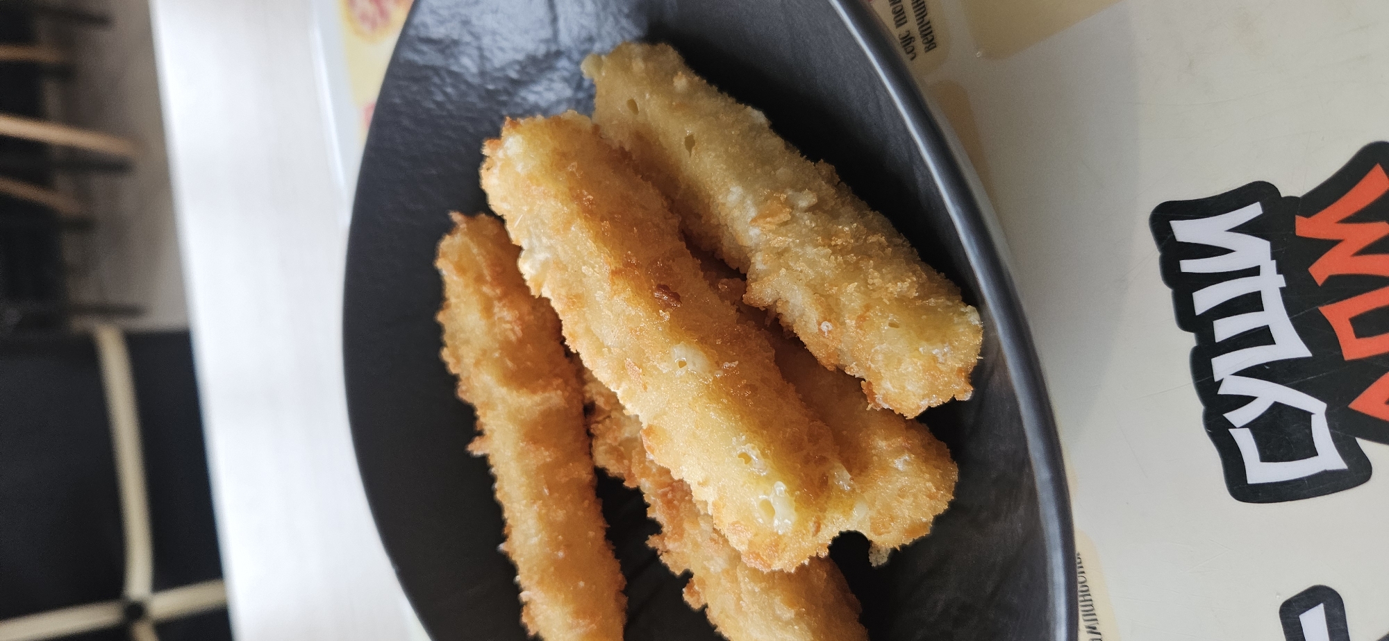 A close-up, top-down shot captures a dark, round bowl filled with several golden-brown, breaded cheese sticks. The cheese sticks are arranged in a slightly overlapping, informal pile, with some showing hints of melted cheese peeking through the crispy coating. The bowl rests on a light-colored, textured surface, possibly a table, with some blurred background elements that suggest an indoor setting, possibly a restaurant. The lighting is soft and natural, highlighting the texture of the fried food. A small portion of a placemat with some text is visible at the bottom right. A sticker with stylized text, appearing to be in Cyrillic script, is partially visible at the bottom. The overall scene is focused on the appetizing presentation of the cheese sticks. The location is the city of Vitsebsk, Belarus, and this image was taken at the Sushi Home restaurant.