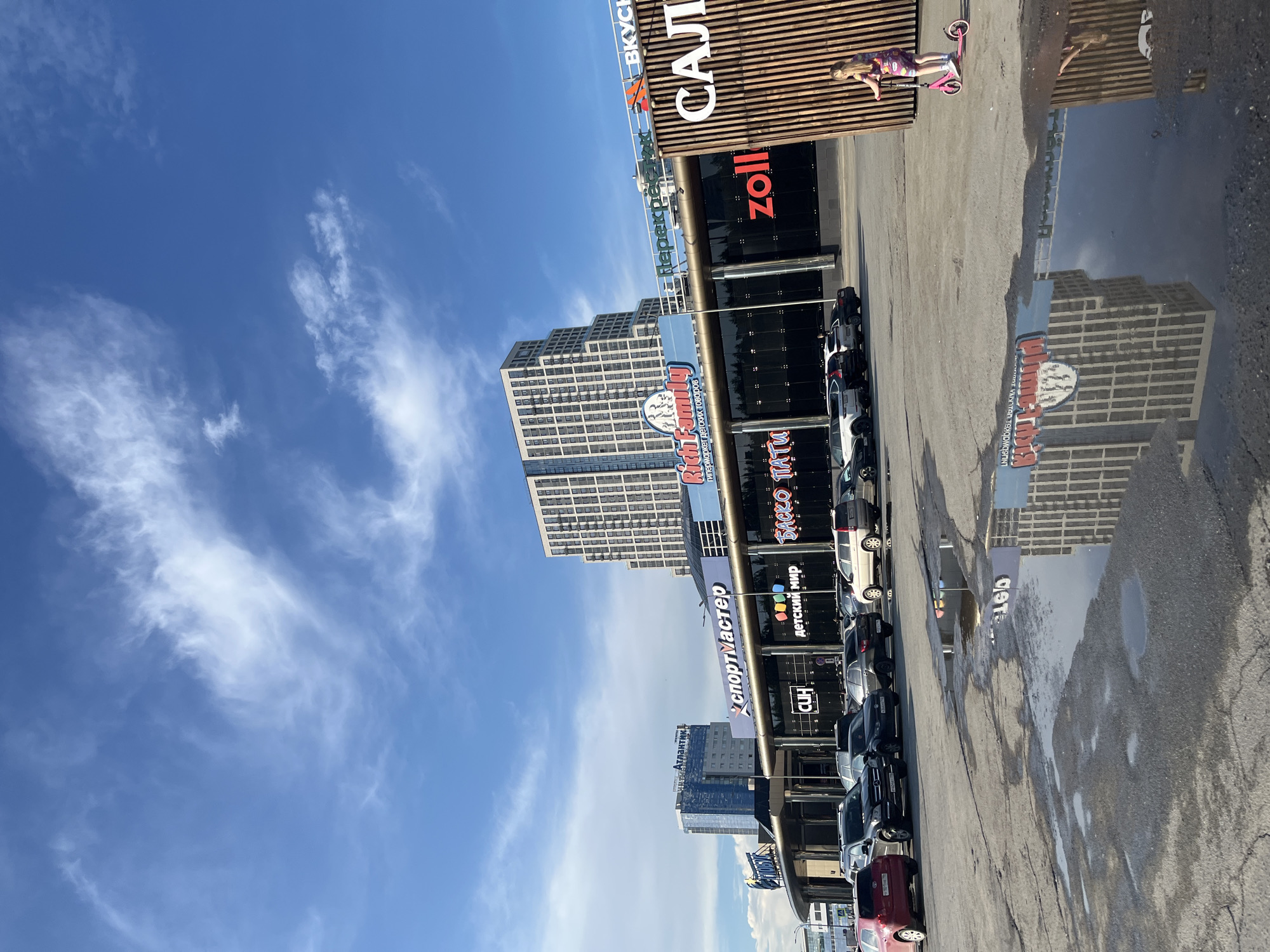 The image depicts a bright, sunny day in Yekaterinburg, Russia, with a clear blue sky dotted with wispy white clouds. The scene focuses on a modern shopping complex, characterized by its sleek architecture and a variety of storefronts with prominent signage.
In the foreground, a parking lot is visible, with several cars parked. Puddles of water on the asphalt reflect the surrounding buildings and the sky. To the right, a young girl on a pink scooter is riding towards the shopping complex.
The shopping complex itself features a long, horizontal building with dark glass facades and illuminated signs. Some of the visible signs include "Sportmaster," "Rich Family" (a children's goods hypermarket), "Detcki Mir" (Children's World), "Zoll," and "Salyut." In the background, taller, multi-story buildings rise, one with a distinct stepped architectural design and another with a blue facade and the "Atlantik" sign.
The overall impression is of a bustling urban environment with a commercial hub, under a clear and pleasant sky. The presence of reflections in the puddles adds a dynamic visual element to the scene.