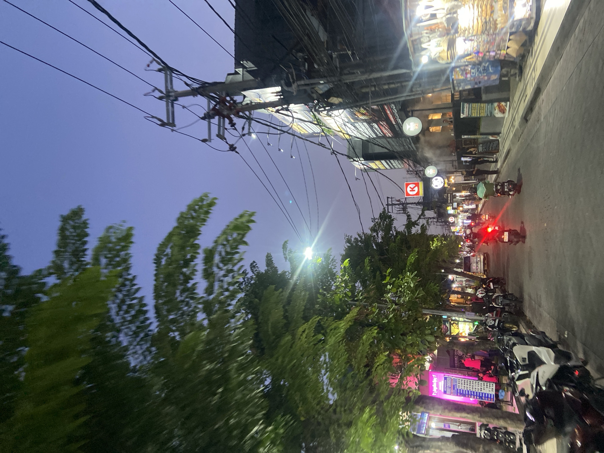 The image captures a lively street scene in Kuta, Indonesia, at dusk or early night, characterized by artificial lights illuminating the thoroughfare and commercial establishments. The perspective is tilted, showing a dense array of overhead utility wires and poles against a deep blue-purple sky on the left. Lush green trees, some appearing blurred from motion, line the street on both sides, with a prominent bright light source causing a lens flare near the center.
On the right side, the street is busy with activity. Numerous motorbikes and scooters are parked along the sidewalk, while others, indicated by red taillights, are in motion. A few pedestrians are visible on the sidewalk. The buildings lining the street house various shops and businesses, displaying illuminated signs and products.
Notable visible text includes a bright pink and purple sign for "REFLEXOLOGY" and "FOOT MASSAGE," which lists prices in Indonesian Rupiah (IDR) for different durations: 10 MIN 20.000, 15 MIN 25.000, 30 MIN 50.000, 45 MIN 75.000, 60 MIN 100.000, 90 MIN 150.000, and 120 MIN 200.000. Other signs include a red square logo with a white "K," which is the "Circle K" convenience store emblem, and round signs featuring stylized coffee cups or similar logos. Another sign partially visible seems to say "BERIK'S" or "ERIK'S." The overall atmosphere is bustling and vibrant, typical of an evening in a tourist-heavy area.