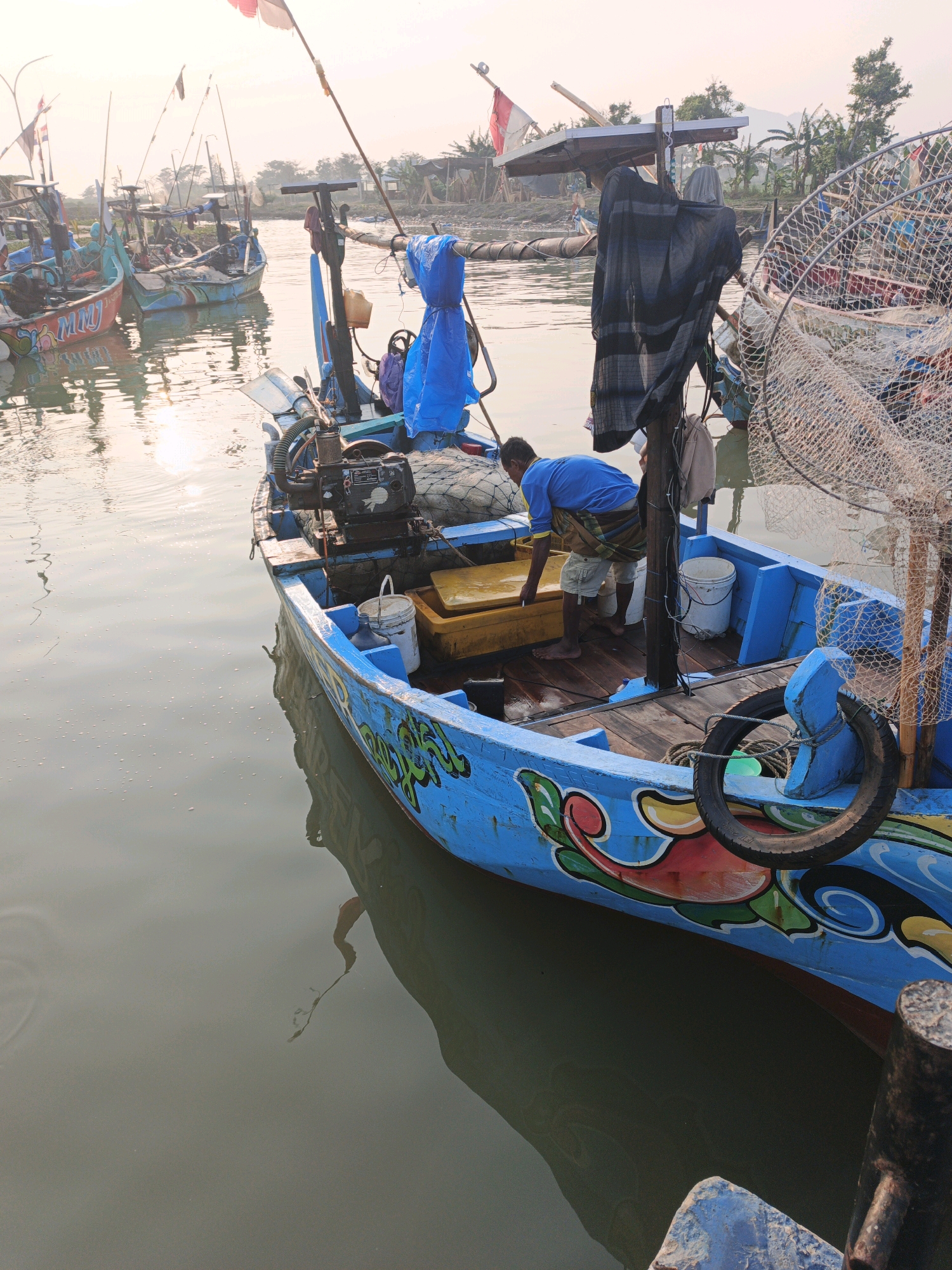 The image depicts a serene waterscape at dawn in the village of Bumiharjo, Indonesia, with several traditional fishing boats docked. The primary focus is on a bright blue fishing boat in the foreground, adorned with colorful artistic motifs along its hull. A man, wearing a blue shirt and checkered sarong, is seen tending to his work on the boat, standing near a yellow storage container. Other boats, painted in various colors and exhibiting unique designs, are visible in the background, partially obscured by mist and the soft morning light. Fishing nets and essential boat equipment, such as an engine and buckets, are also present. The water is calm, reflecting the warm hues of the sunrise and the surrounding scenery. The overall atmosphere is peaceful, suggesting the start of a typical day for the fishing community in Bumiharjo.