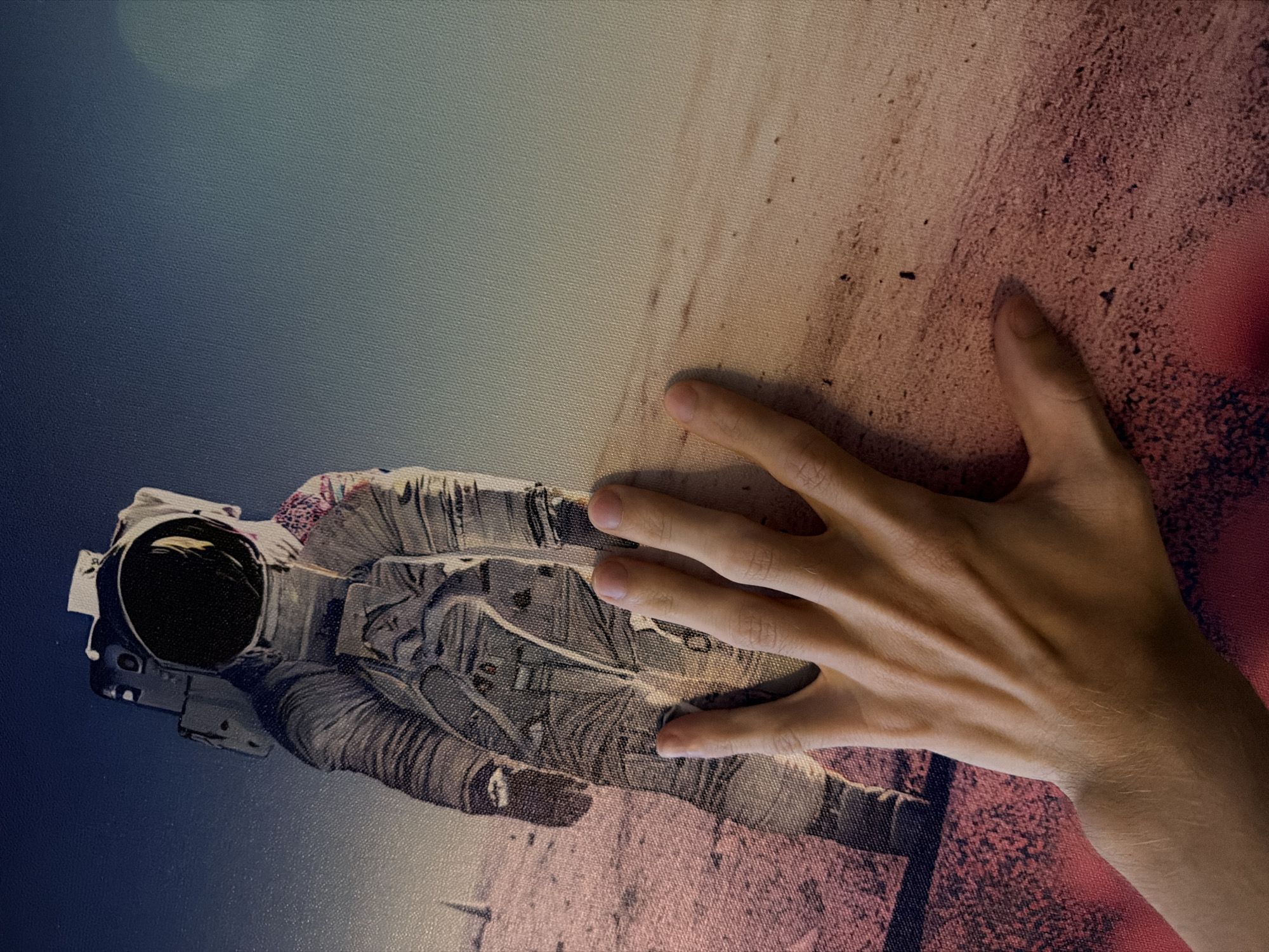 The image shows a close-up of a hand resting on a large canvas print. The print depicts an astronaut standing on a barren, dusty landscape with a gradient sky. The astronaut is positioned in the upper left quadrant of the image, while the hand occupies the lower half and extends towards the right. The hand is light-skinned, with visible veins and fine hairs on the forearm. The fingers are slightly splayed, with the thumb and index finger positioned near the astronaut's suit, and the other fingers resting on the sandy terrain. The texture of the canvas is visible throughout the image. There is no discernible connection to the city of Naberezhnye Chelny, Russia.