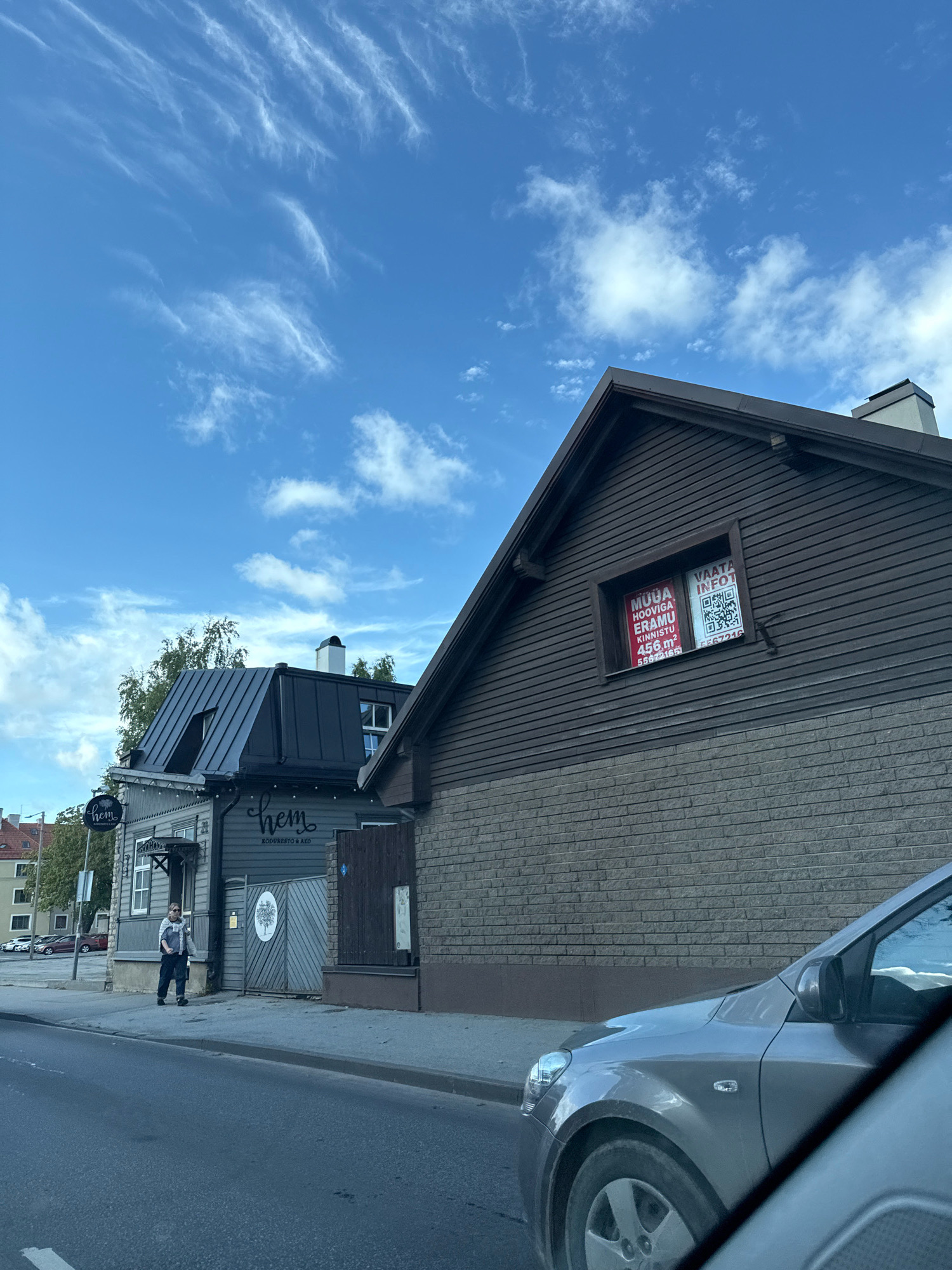 The image captures a street scene in Tallinn, Estonia, under a partially cloudy blue sky. In the foreground, a silver car is partially visible, obscuring the lower right portion of the frame. The middle ground features a row of buildings. The most prominent building on the right is a dark brown wooden structure with a pitched roof and a brick-clad lower section. A window on the upper part of this building displays two signs: a red and white sign with text and a QR code, and a white sign with a QR code. To the left of this building is a narrower, darker building with a darker pitched roof and an entrance. A sign reading "hem" is visible on this building, along with text below it that appears to say "KODURESTO & ARO". A person in casual clothing, including a jacket and dark pants, is standing on the sidewalk in front of this building, facing left and holding a bag. Further to the left, other buildings and parked cars are visible in the distance. The street in front of the buildings is paved with asphalt.