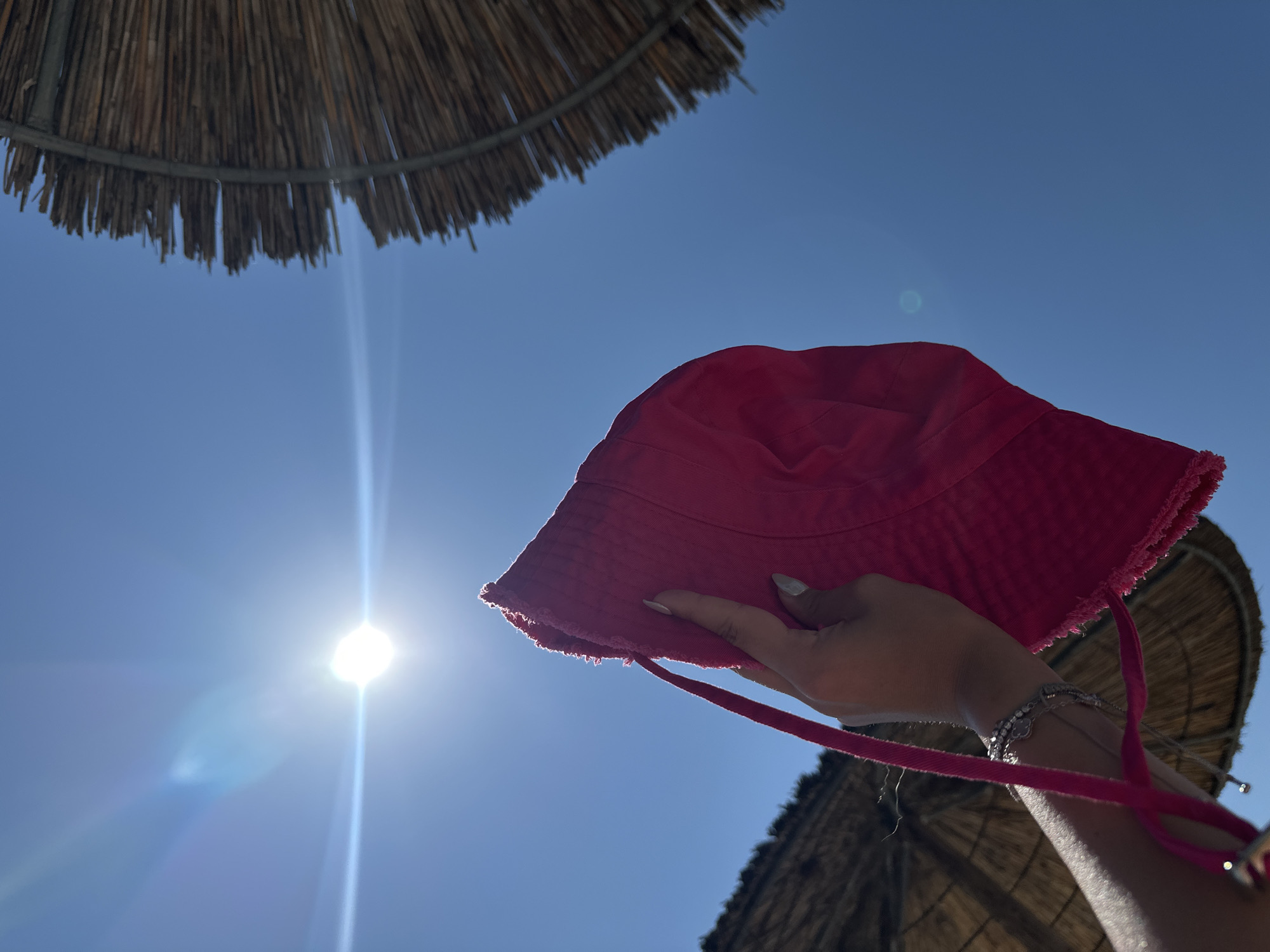 A person's hand holds a bright pink bucket hat against a clear blue sky. The sun is high and bright, causing lens flare. Two thatched beach umbrellas are visible in the background, one on the left and one on the right. The image appears to have been taken at Ofrynio Beach, Greece, as indicated by the context. The scene is outdoors, likely on a sunny day at the beach.