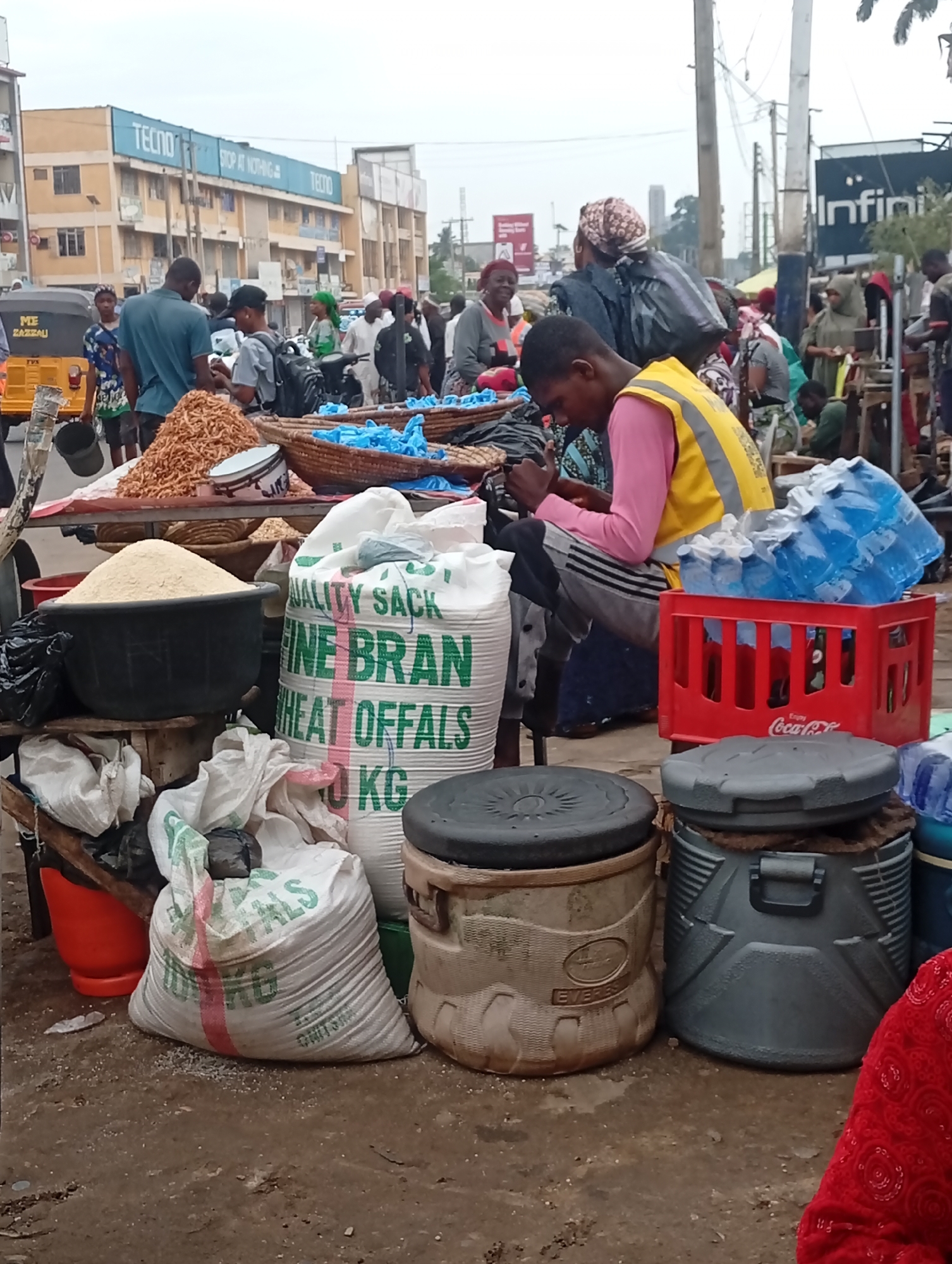 The image depicts a bustling street scene in Maiburiji, Nigeria. A vendor wearing a yellow vest and pink shirt sits on a stool, appearing to be using a mobile phone. He is surrounded by goods for sale, including bags of "FINE BRAN WHEAT OFFALS," containers of what appears to be rice or flour, dried shrimp, and bottles of water stacked in a red Coca-Cola crate. Other people are visible in the background, along with a "TECNO" sign on a building and a yellow tricycle labeled "ME ZAZZALU." The ground appears to be wet and dirty.