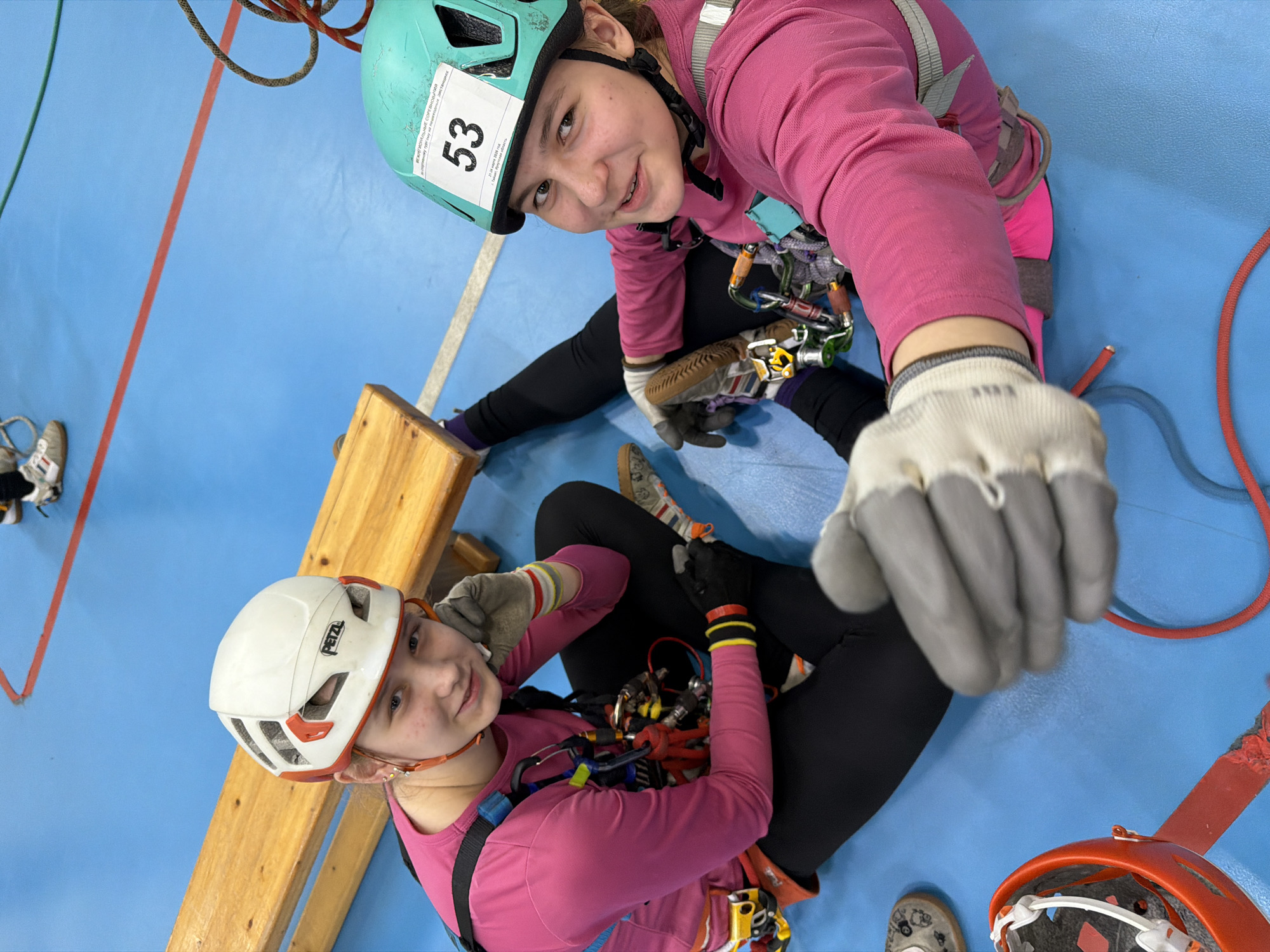 Two young people wearing climbing helmets and harnesses are positioned close to each other against a blue background with red and orange lines. The person at the top of the frame is wearing a teal helmet with the number "53" on it, a pink long-sleeved shirt, black leggings, and climbing shoes. They are extending their gloved hand towards the viewer. The second person, positioned lower in the frame, is wearing a white Petzl helmet, a pink long-sleeved shirt, black leggings, and climbing gloves. They are sitting next to a wooden structure. Climbing equipment and ropes are visible. The scene appears to be indoors, potentially at a climbing gym or training facility. No location information is visibly apparent.