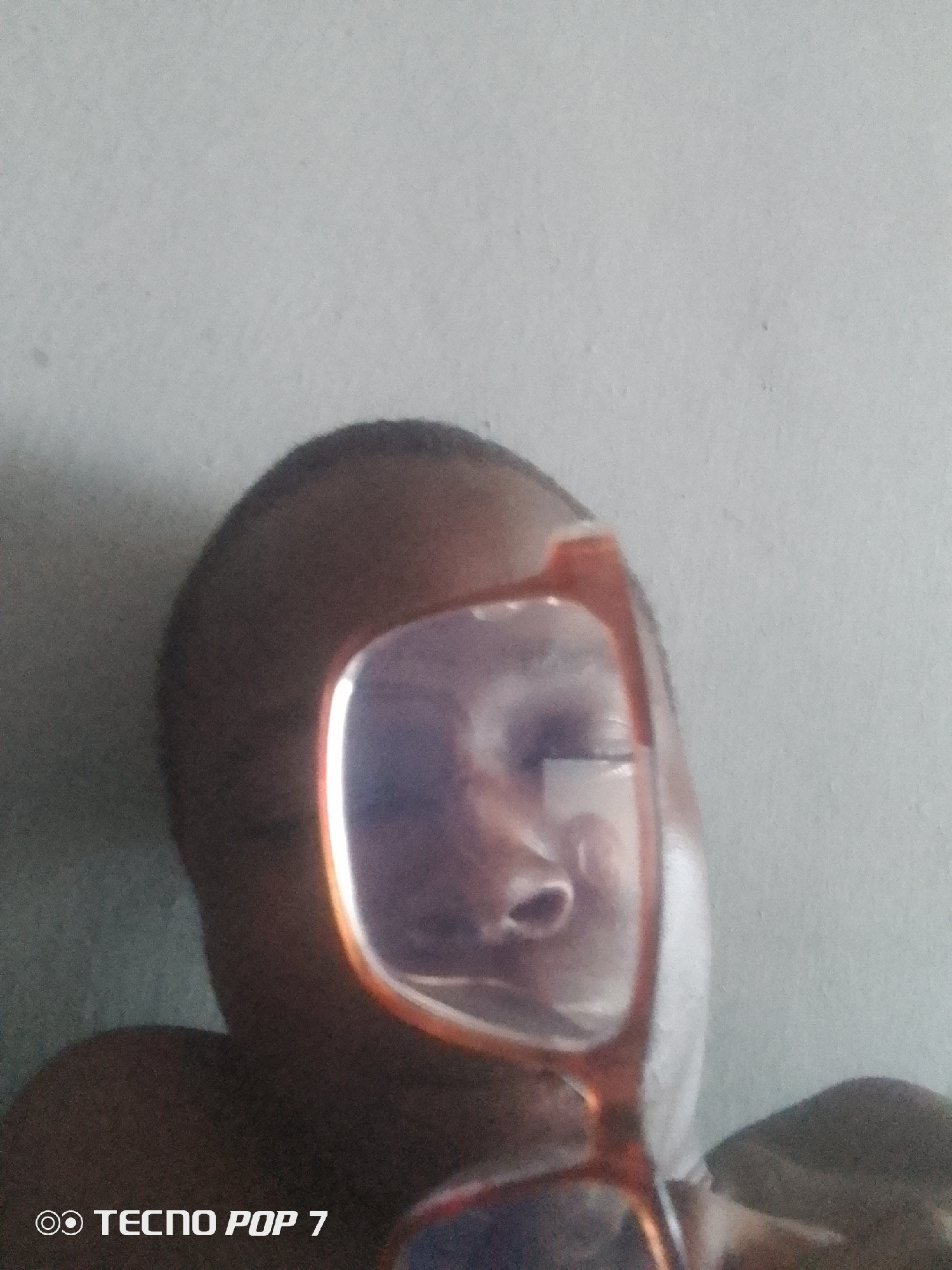 A close-up, overhead shot shows a person's head, mostly obscured by a pair of orange-rimmed glasses. The glasses are positioned in front of the person's face, and only the eyes and nose are visible through the lenses. The person's skin tone is dark, and their hair is short and dark. The background is a plain, light-colored wall. A watermark in the bottom left corner reads "TECNO POP 7". The setting appears to be indoors, in Ipetumodu, Nigeria.