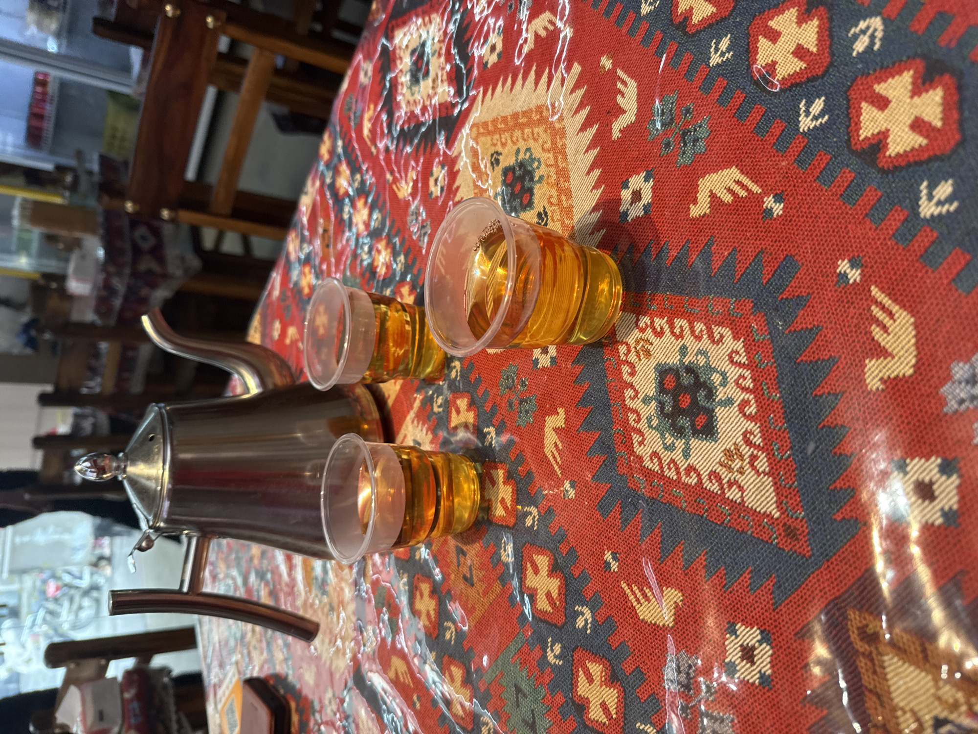 A close-up shot captures a metal teapot and three small plastic cups filled with amber liquid, likely tea, arranged on a table covered with a red, blue, and beige patterned tablecloth. The tablecloth features geometric designs and stylized animal motifs, reminiscent of traditional Middle Eastern or Central Asian textiles. The teapot is centrally located, with the cups spaced out in front of it. The lighting is warm and indoor, suggesting a cozy dining or cafe setting. The background is blurred, but hints of wooden chairs and tables, as well as what appears to be a shelf with various items, are discernible, suggesting an indoor dining establishment. The image does not provide any specific visual cues to confirm its location in Huizhou, China.