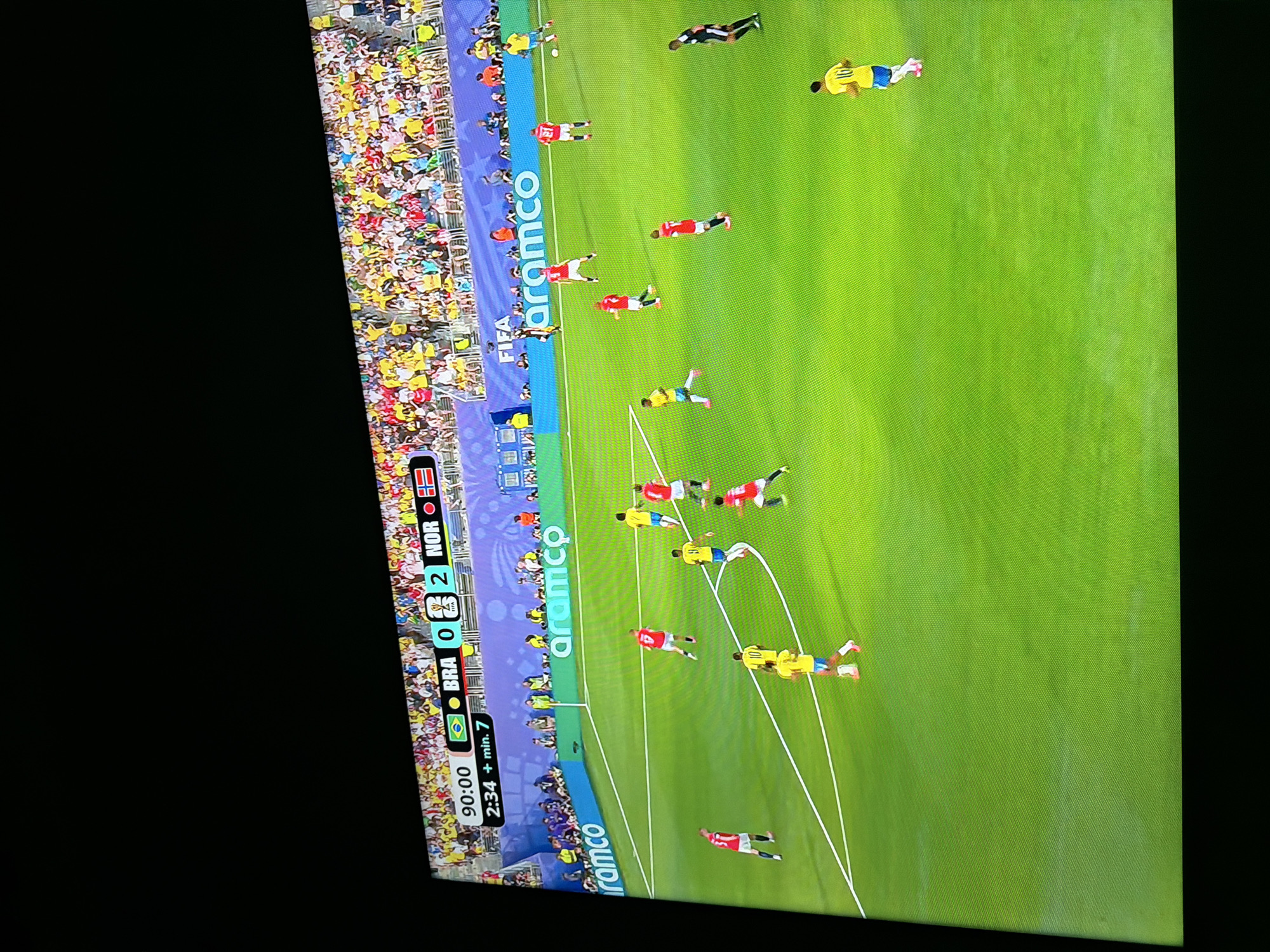 This is a photograph of a soccer match being broadcast on a television screen. The score displayed at the top of the screen shows Brazil 0 and Norway 2. The game clock indicates 90:00, with an additional 2 minutes and 7 seconds of stoppage time. The field is green with white markings, and several players in red and yellow uniforms are visible. A crowd is present in the stands behind the field. Advertisements for "Aramco" are displayed prominently on the stadium walls. There is no discernible indication of the city of Ognianovo, Bulgaria, in this image.